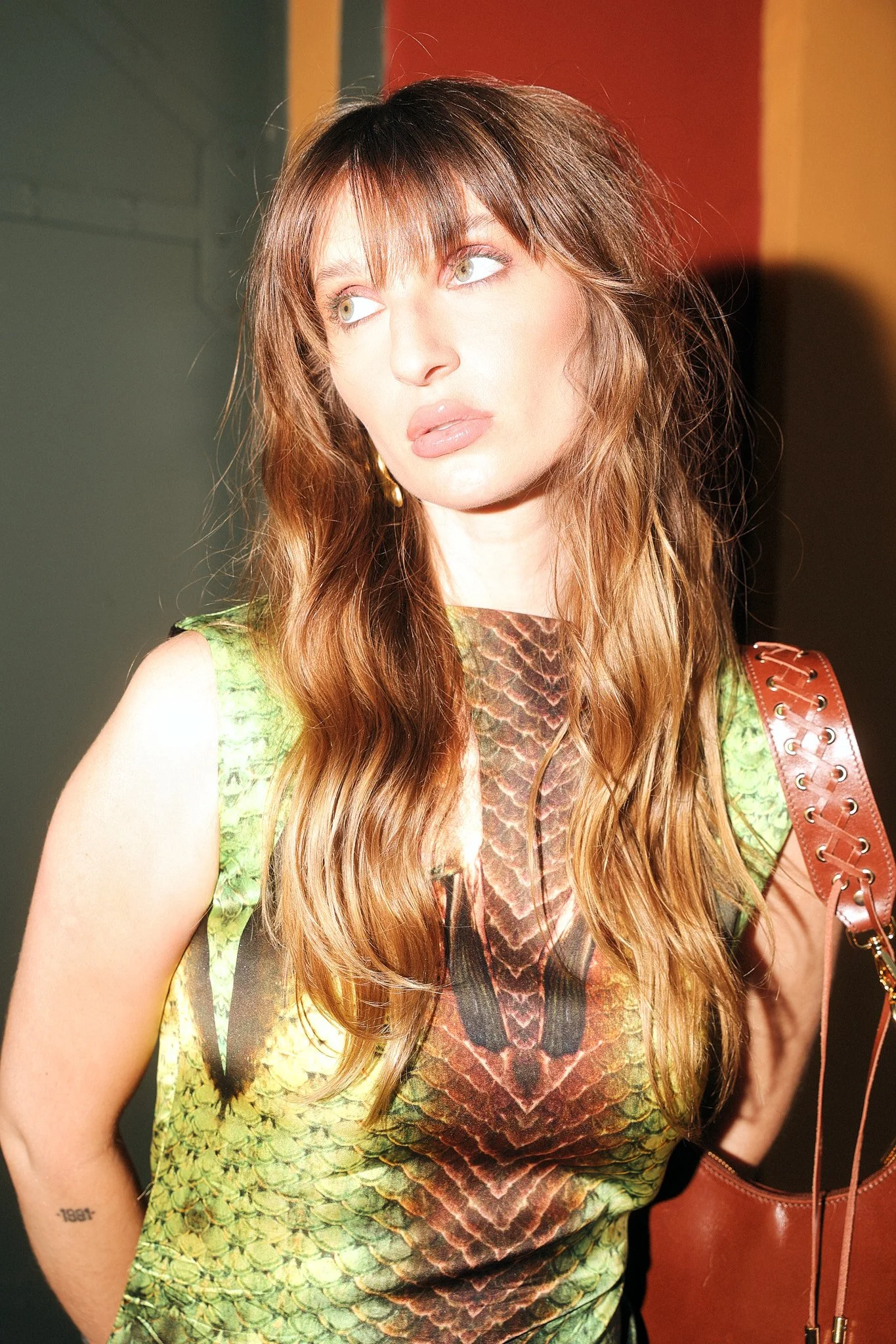 Mulher com cabelo longo, castanho claro e olhos verdes, vestindo uma blusa com estampa de cobra e carregando uma bolsa marrom com detalhes de metal.