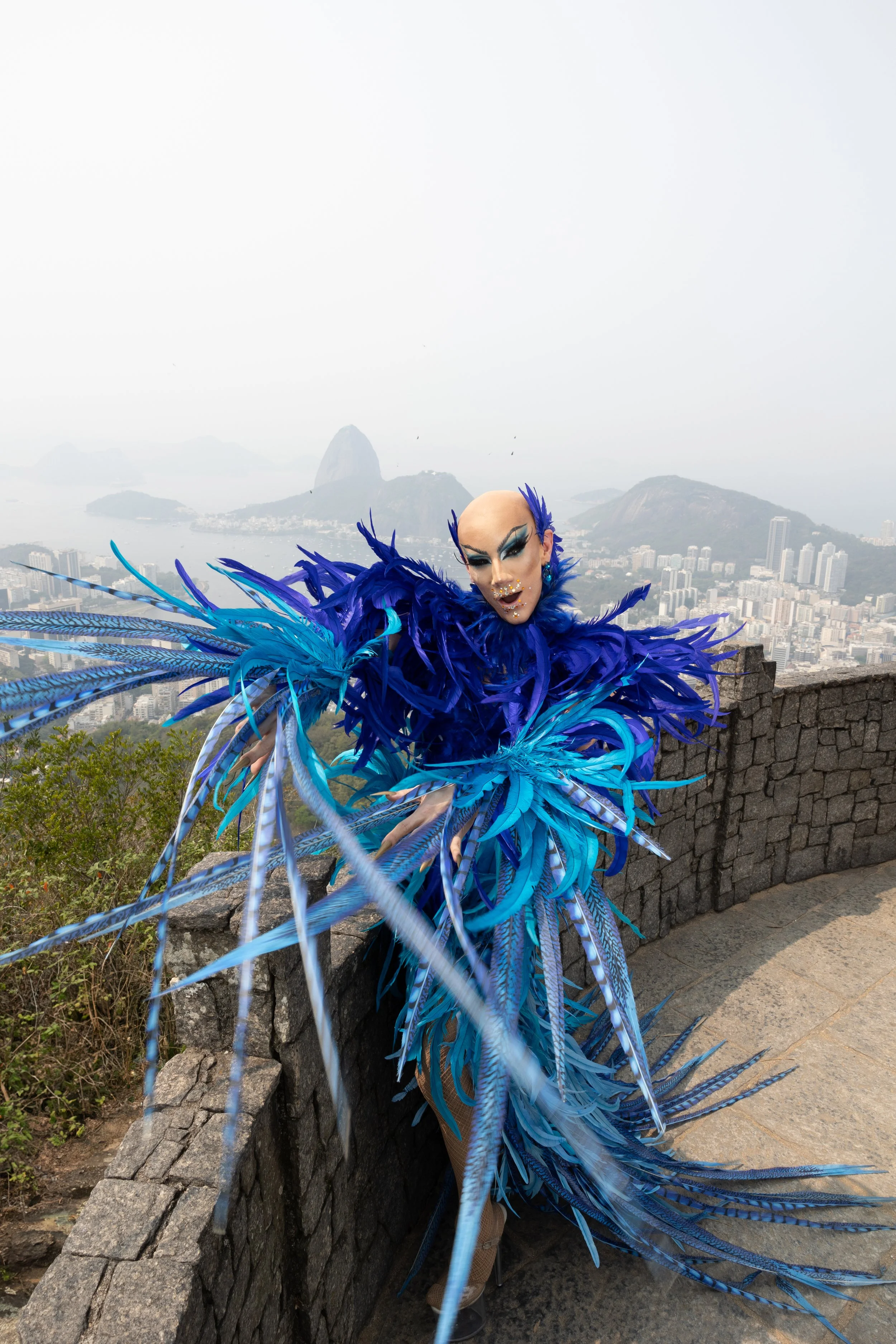 Pessoa vestida com roupa de penas em tons de azul, com maquiagem teatral, posando em local elevado com vista de cidade e montanhas ao fundo.