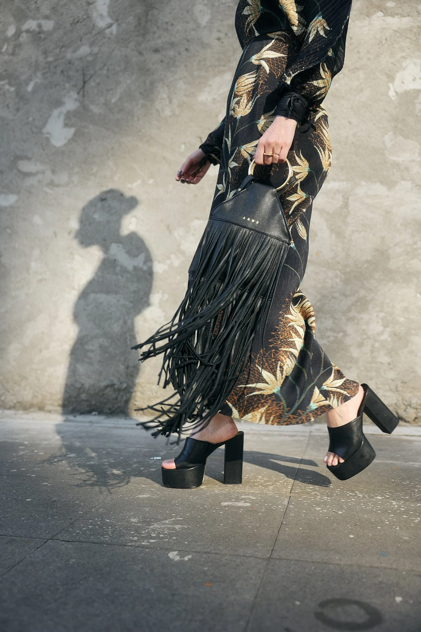 Pessoa usando sapatos de salto grosso preto, saia ou vestido com estampa floral, carregando uma bolsa preta com franjas e um anel na mão, caminhando ao ar livre com uma parede de fundo de concreto.