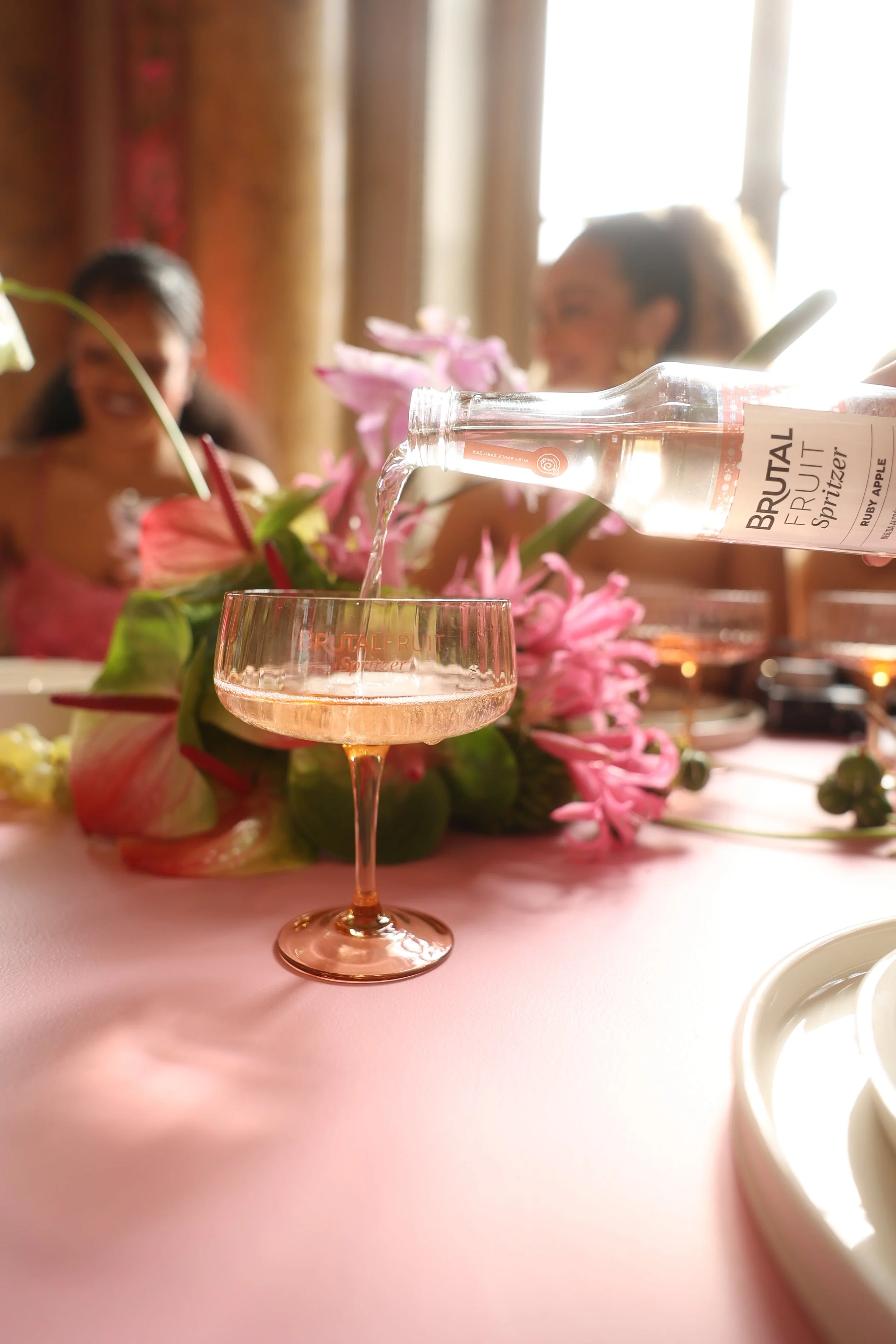 Garrafa de água de sabor de fruta ruiva sendo despejada em uma taça de champagne com uma mesa decorada com flores ao fundo.