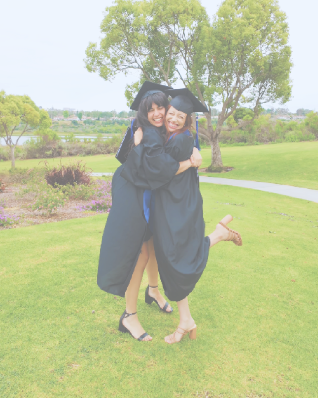 Two women in graduation cap and gown hugging and smiling outdoors on a grassy area with trees and a river in the background.