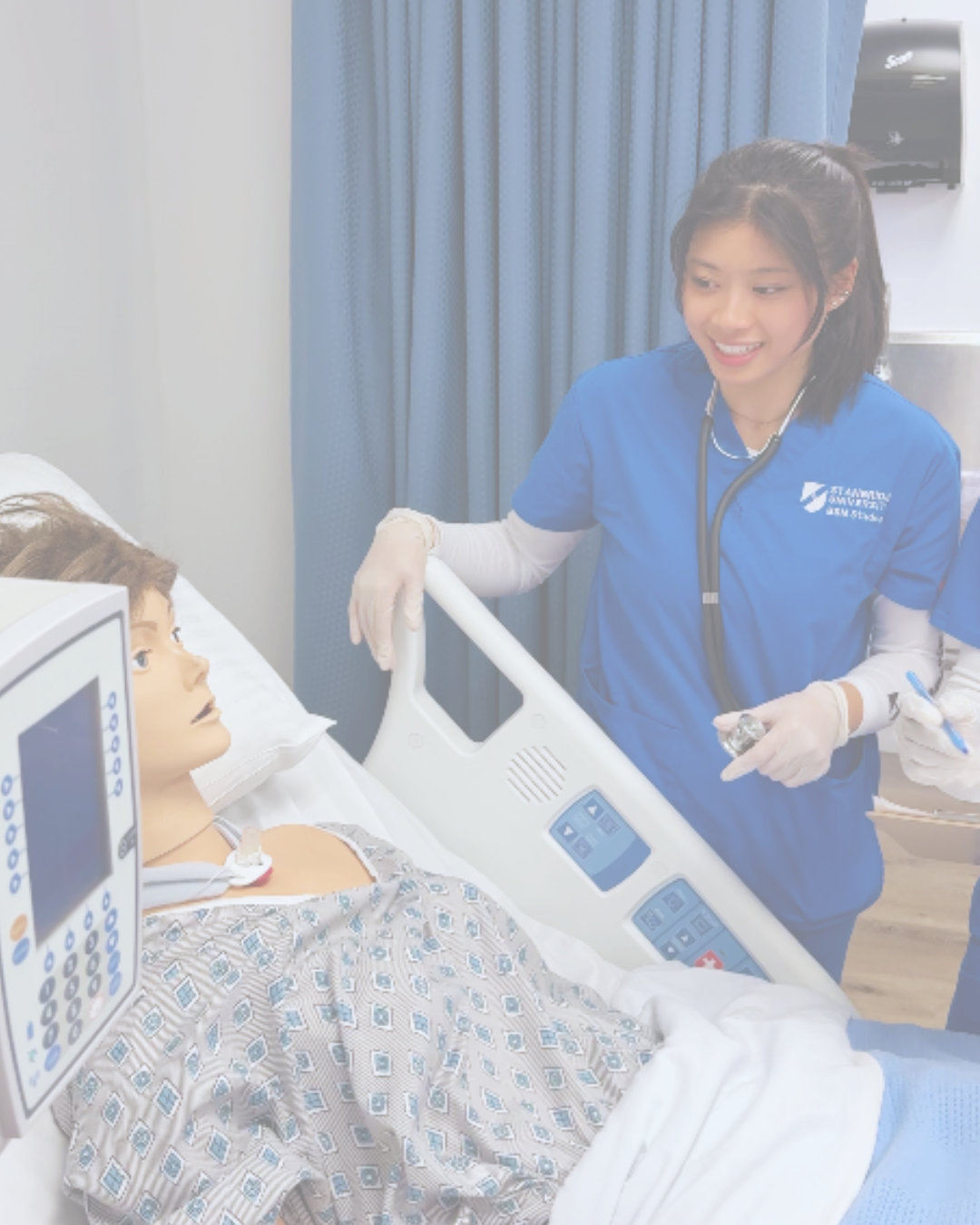 A nurse in blue scrubs and gloves smiling and talking to a patient lying in a hospital bed.