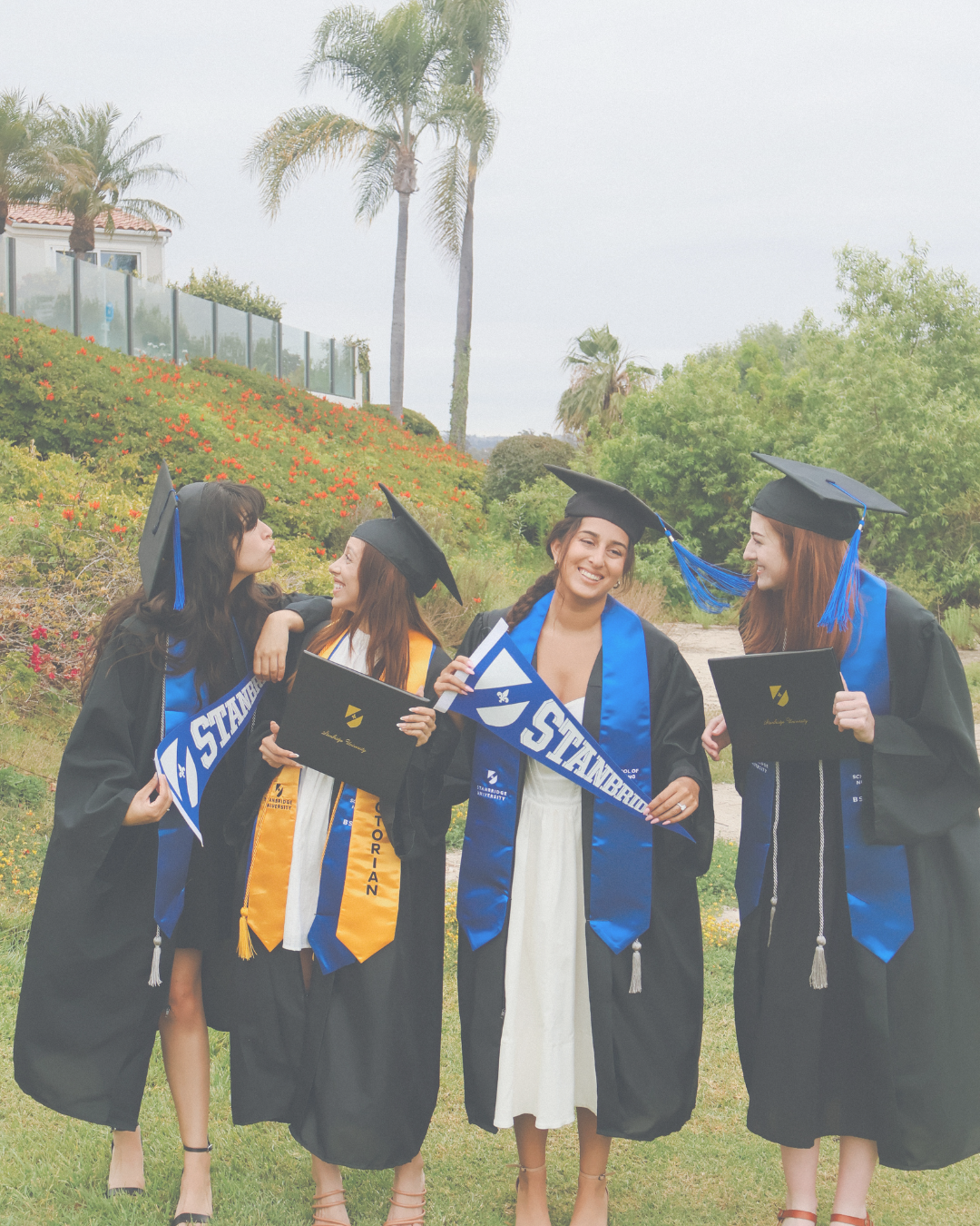 Four women in graduation caps and gowns celebrating outdoors, holding diplomas and college flags, with greenery and palm trees in the background.