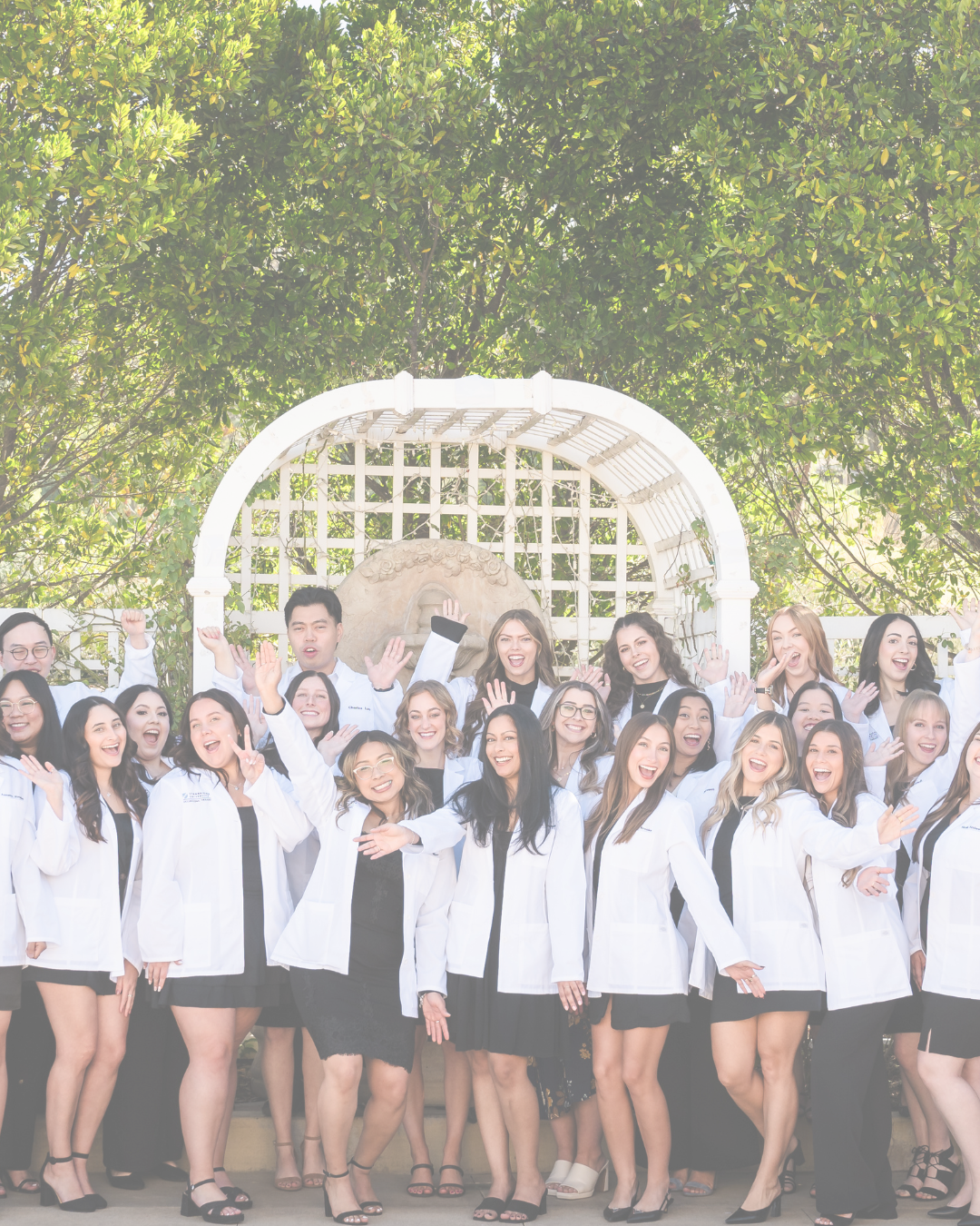 Group of smiling women and men in white coats celebrating outdoors in front of a garden archway.
