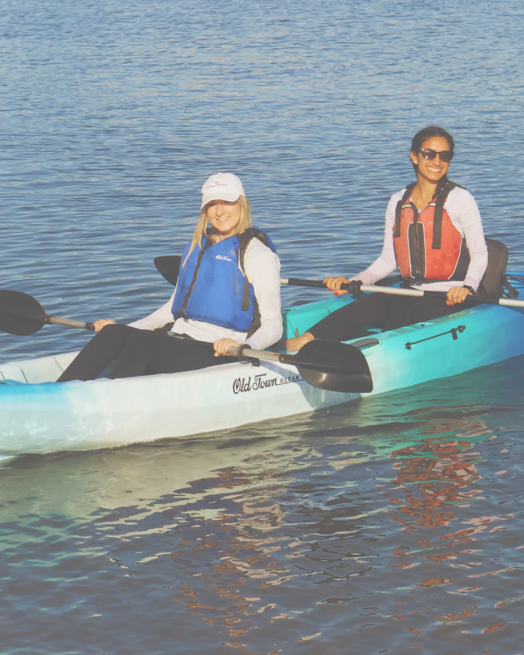 Two women kayaking on calm water, wearing life jackets and sunglasses, smiling at the camera.