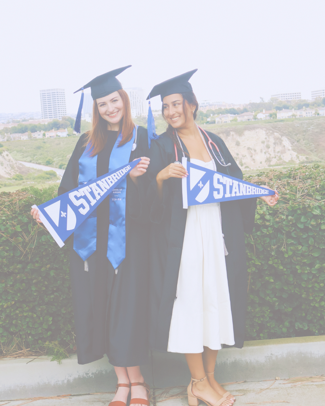 Two women in graduation gowns and caps holding Stanford University pennants, standing outdoors with city and greenery in the background.