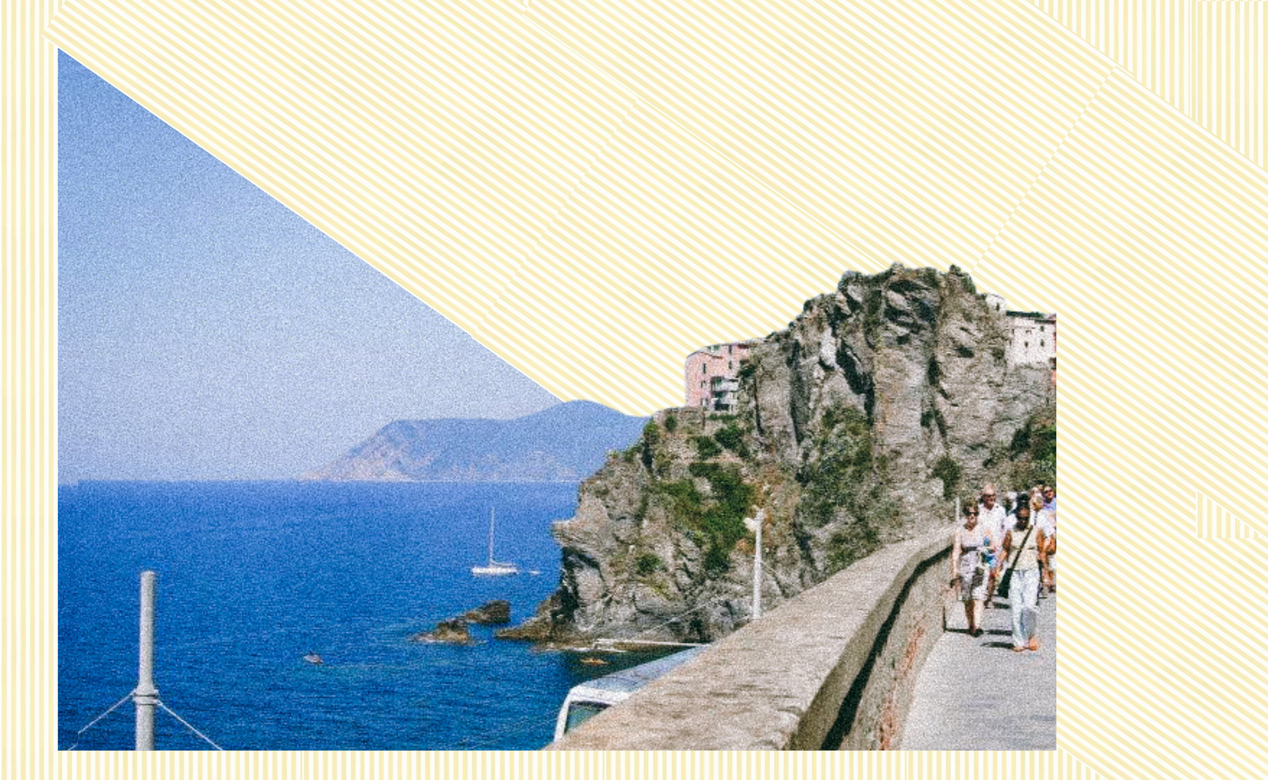 Tourists walking along a coastal pathway on a rocky cliffside overlooking the sea, with mountains in the background and sailboats on the water.