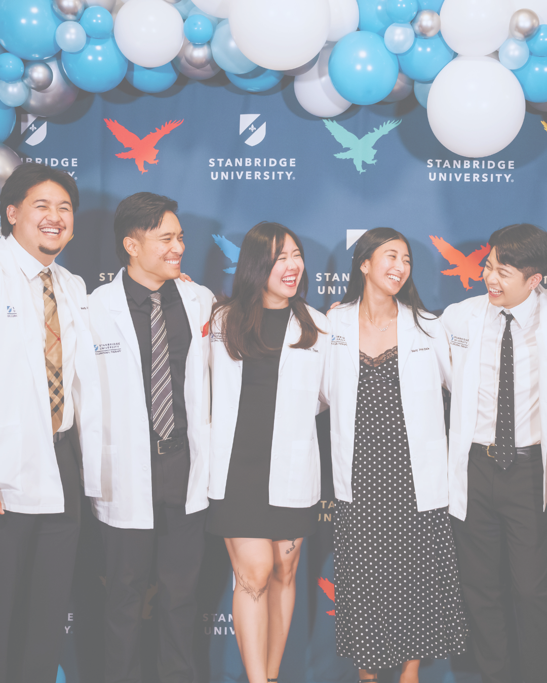 Group of five students in white coats smiling and laughing at graduation or academic event, standing in front of a blue backdrop with balloons and the Stanford University logo.