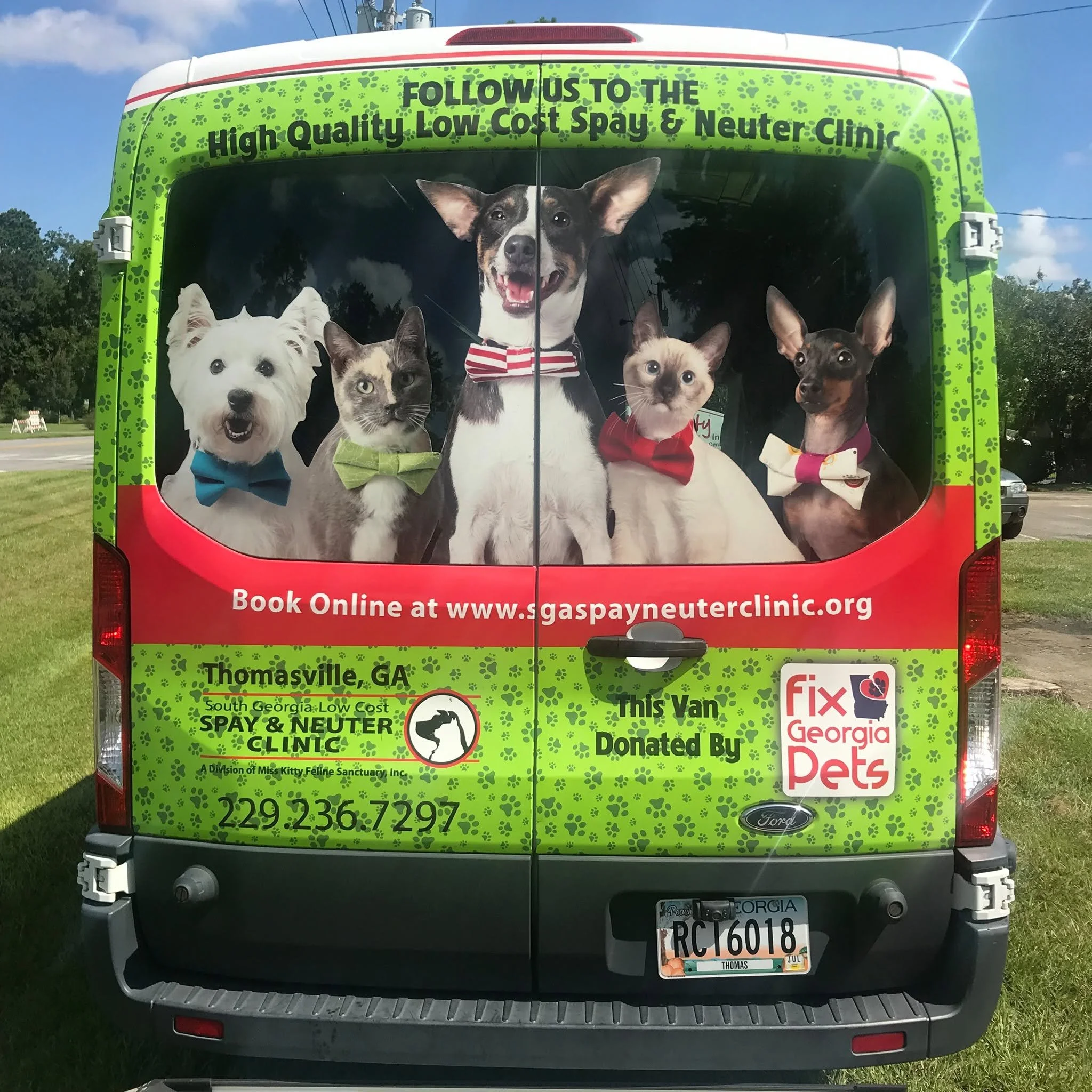 The back of a colorful van with a large graphic of five pets dressed in bow ties and wearing accessories. The van advertises a pet clinic in Thomasville, Georgia, specializing in low-cost spay and neuter services. The text on the van includes a website and phone number for booking appointments.