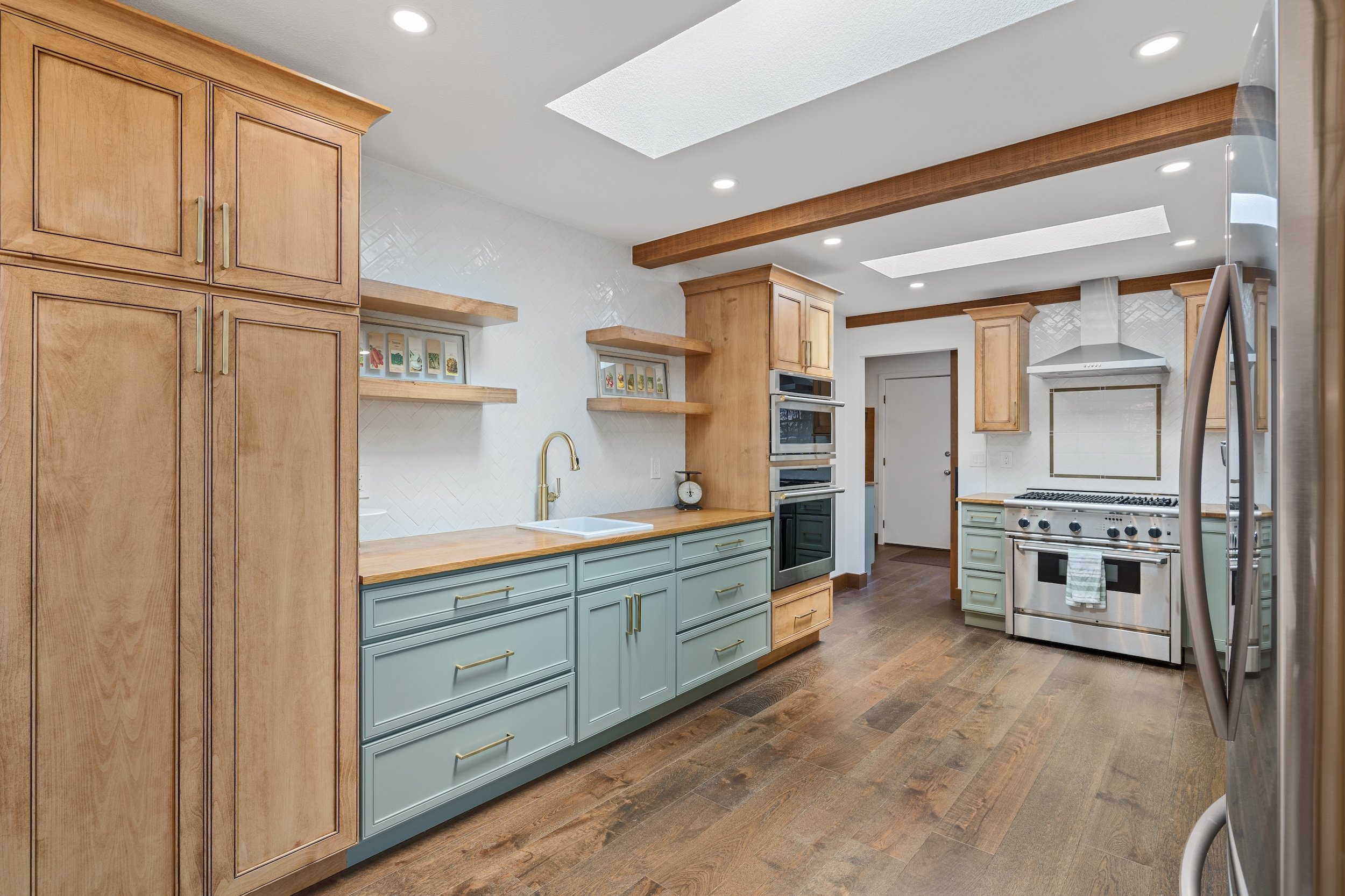 A modern kitchen with wooden cabinets, light blue lower cabinets, and stainless steel appliances. Wooden beams on the ceiling, recessed lighting, and a white tiled wall with shelves displaying framed pictures. Wooden flooring and a stainless steel re