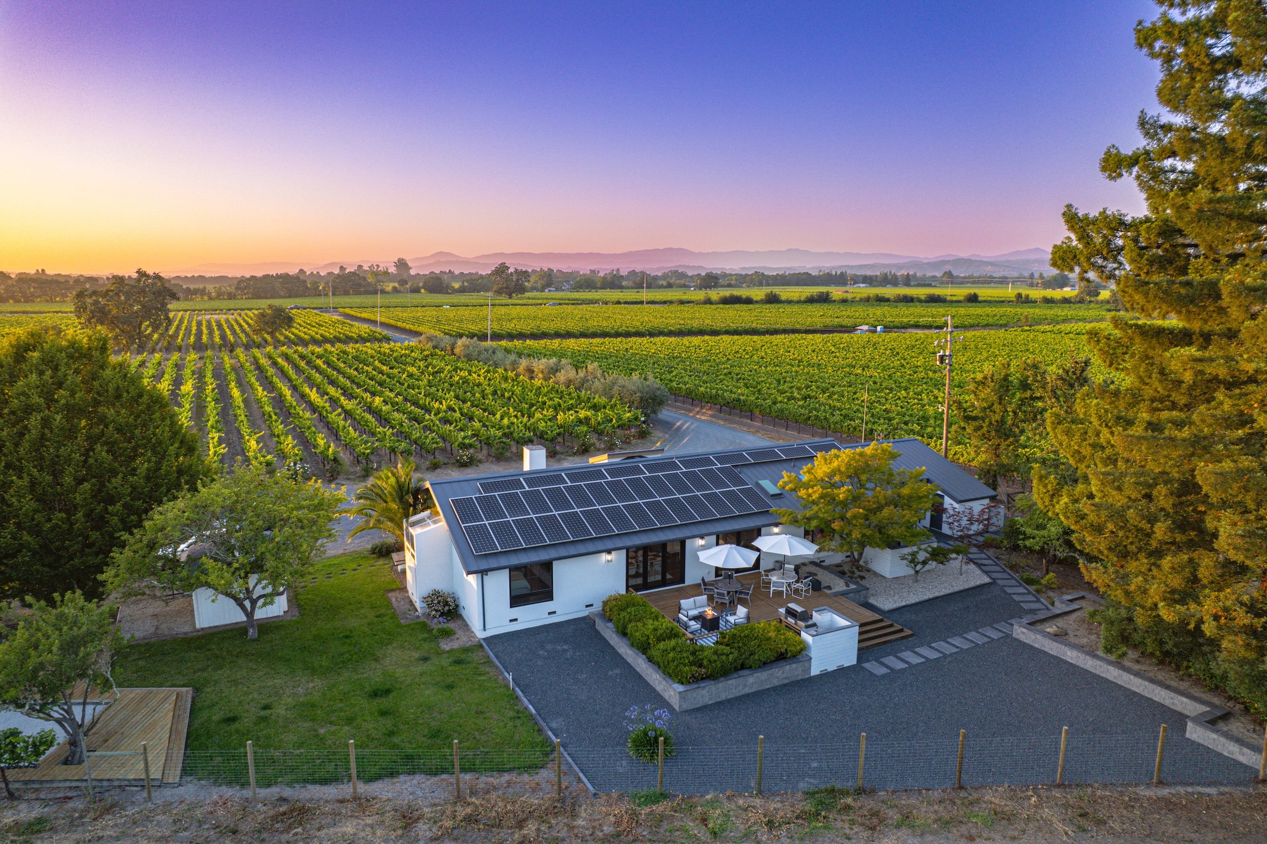 A house with solar panels on the roof amidst a lush landscape with vineyards and trees, with mountains and colorful sunset sky in the background.