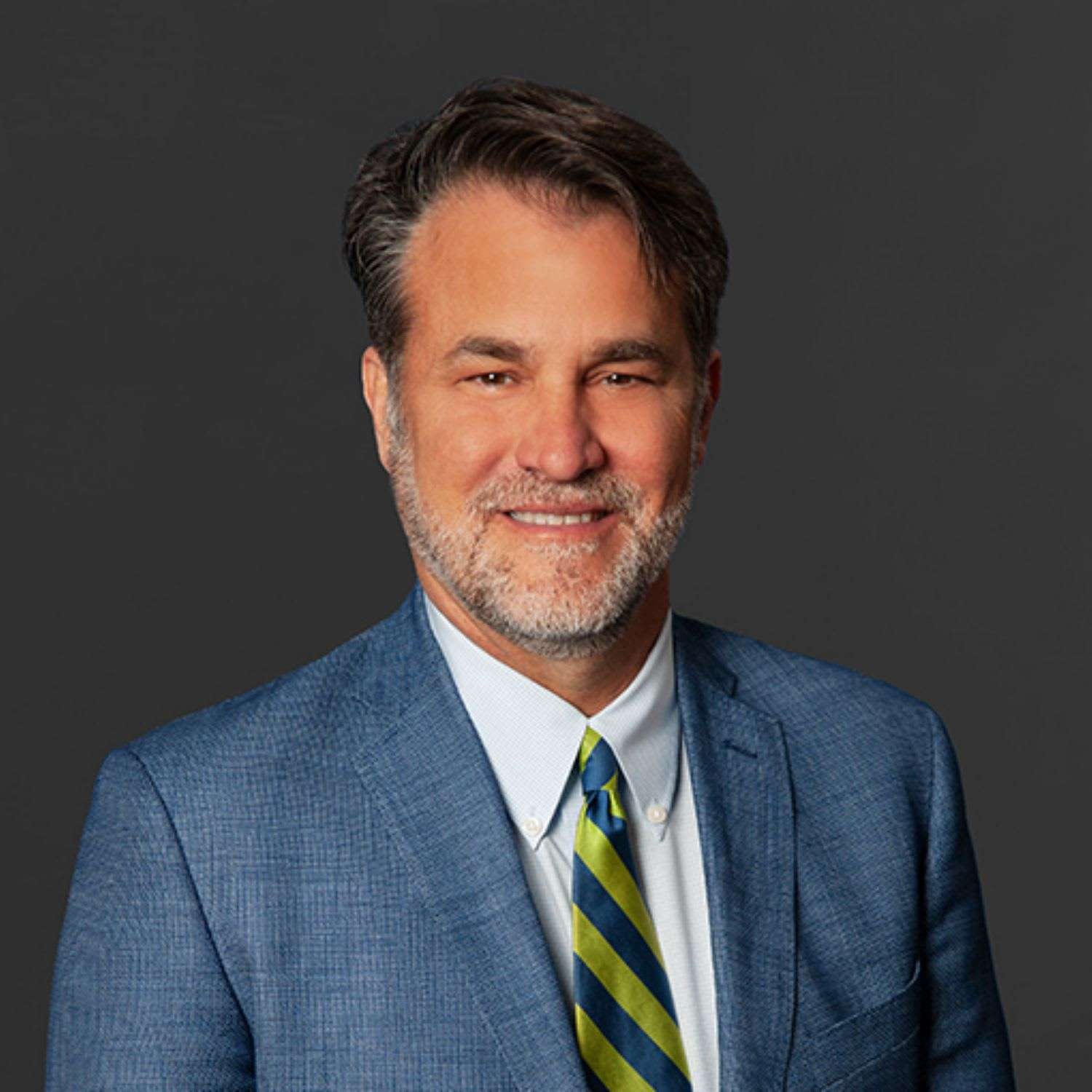 Well-dressed middle-aged man with dark hair and gray beard, smiling, wearing a blue suit, white shirt, and a striped tie, against a dark gray background.