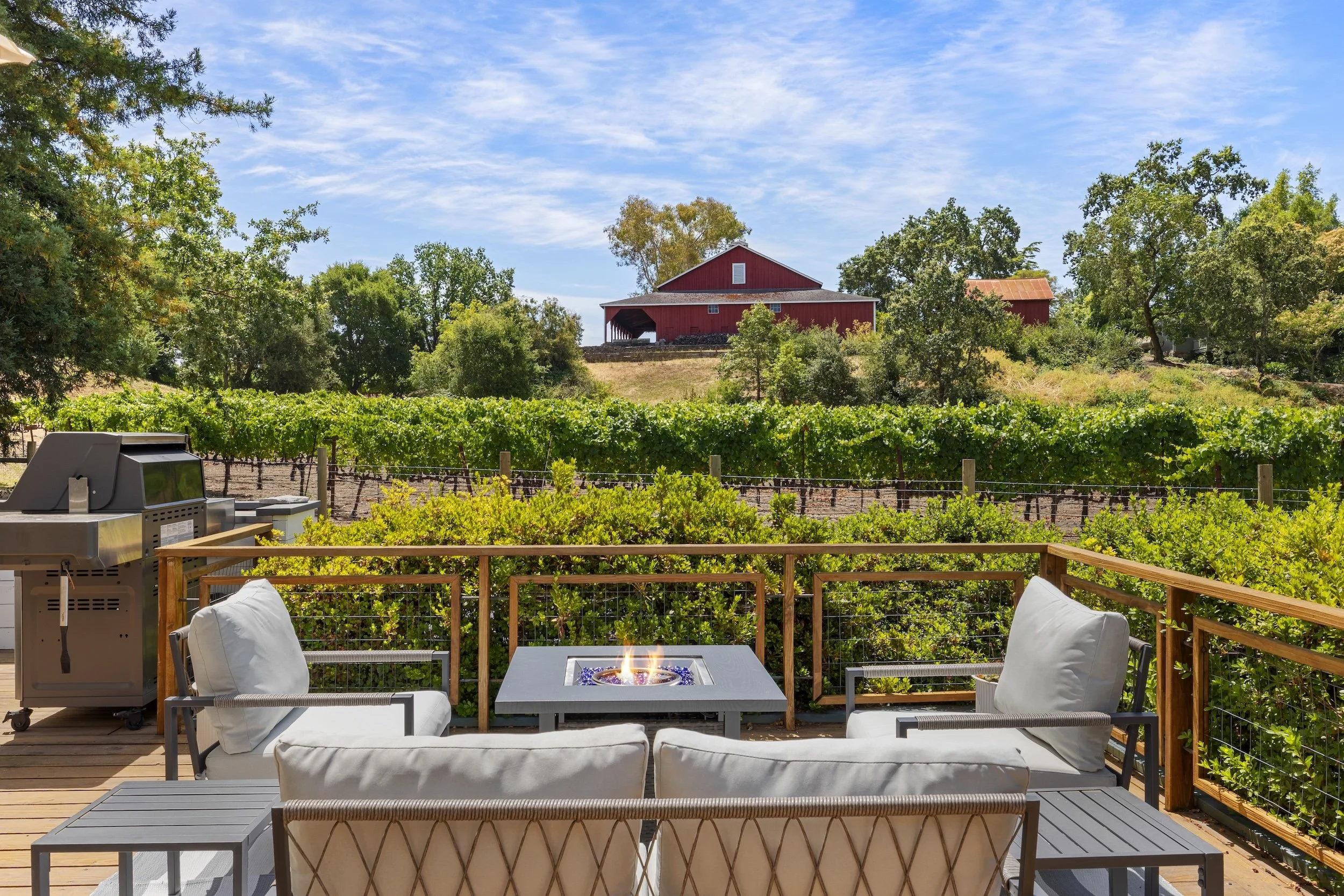 A backyard patio with white cushioned chairs around a fire pit, overlooking a vineyard and a red barn surrounded by trees and a blue sky.