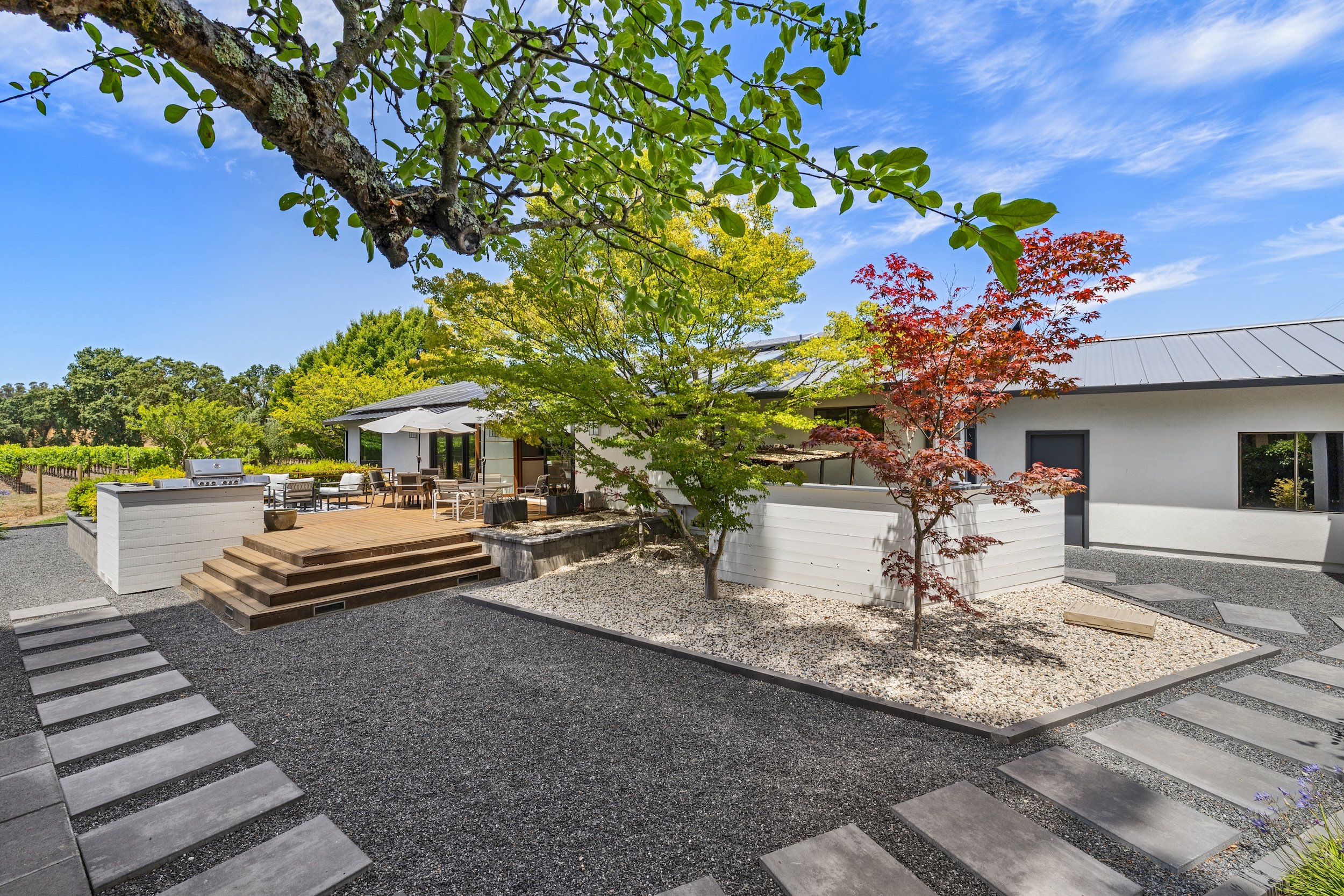Modern house with white exterior walls, a metal roof, and a spacious outdoor patio area with seating, surrounded by landscaped garden beds with trees and steps leading to the patio.