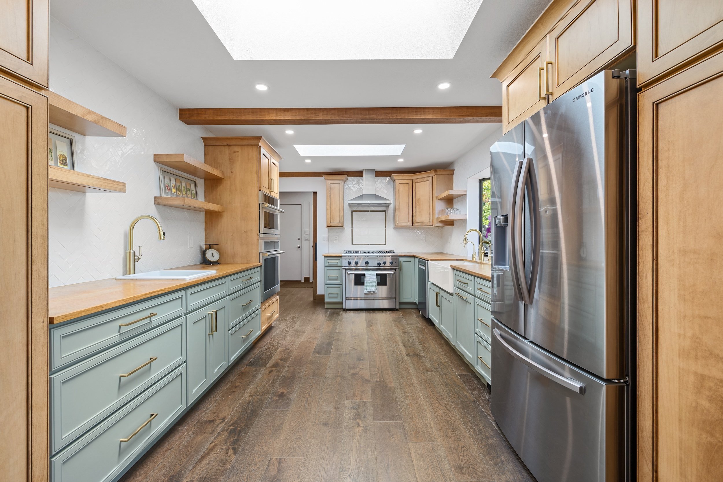 A spacious kitchen with wooden cabinetry, stainless steel appliances, and a large sink with a gold faucet. Light blue cabinets are complemented by natural wood countertops and open wooden shelves. Skylights and recessed lighting illuminate the room, 