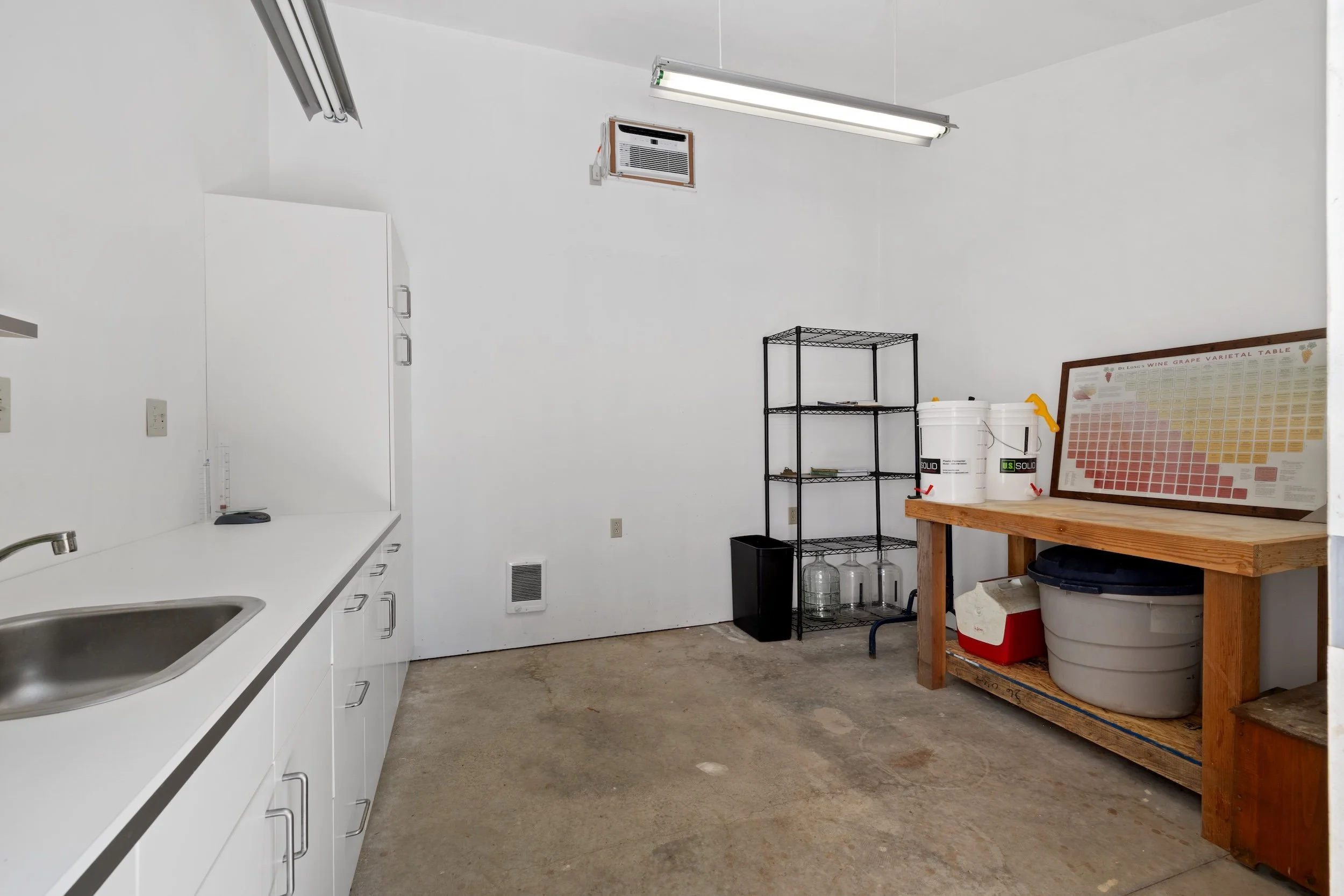 A clean, minimalist kitchen with a white countertop and cabinetry on the left, a wall-mounted air conditioner, a fluorescent ceiling light, a black metal shelving unit with water containers, a framed wine grape chart, and a wooden table with storage 