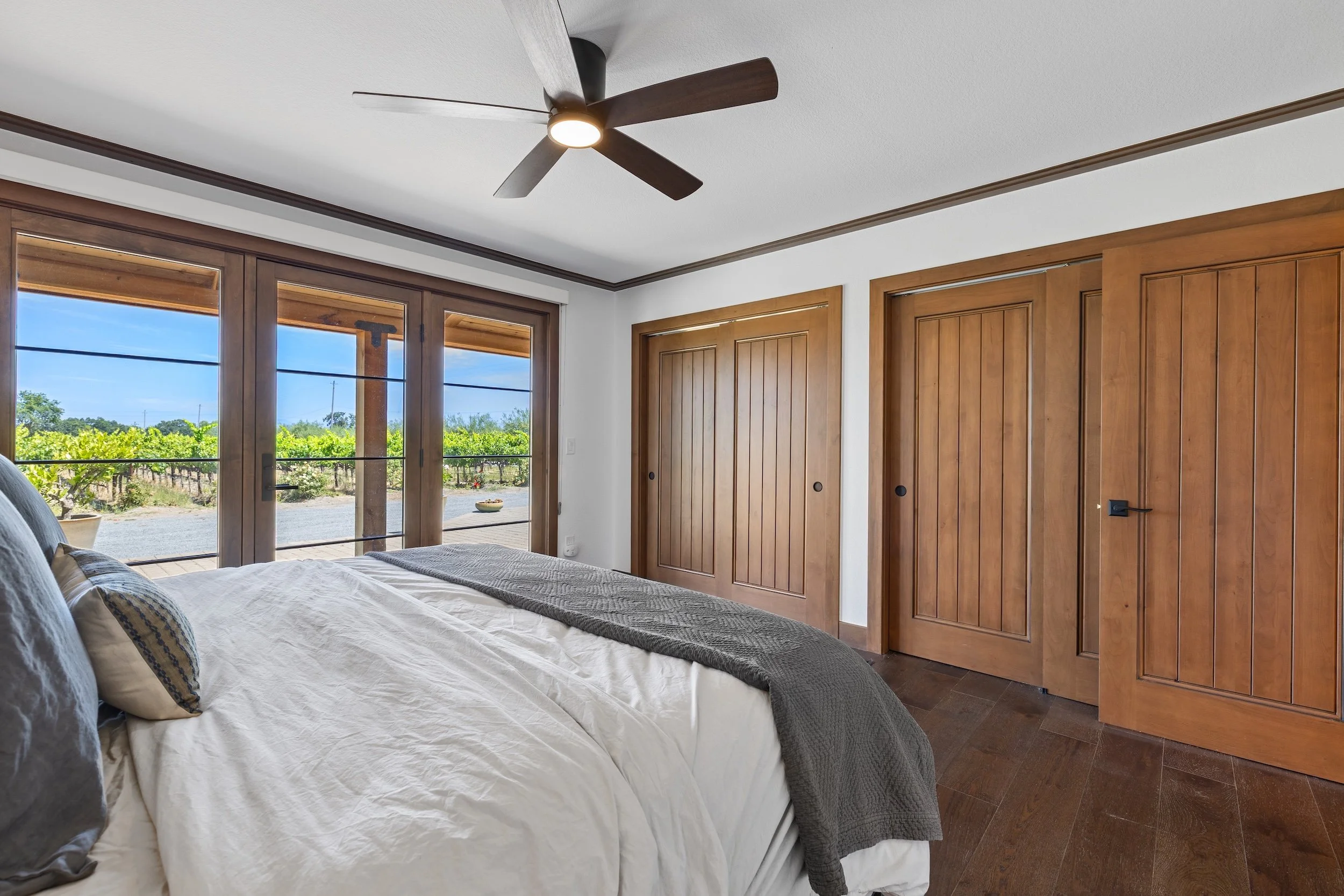 Bedroom with white walls, wooden closet doors, large windows showing a view of vines outside, bed with white bedding and pillows, ceiling fan with dark blades.