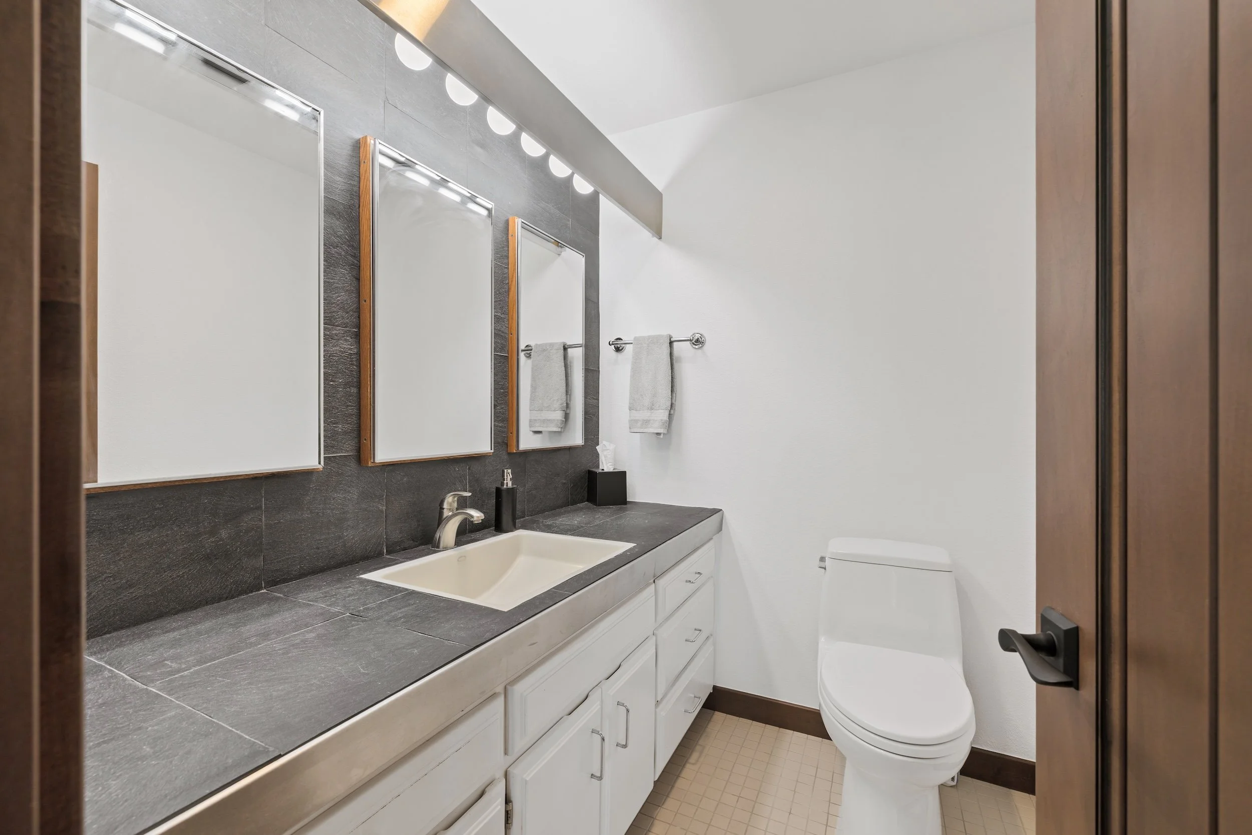 A modern bathroom with dark gray tiled wall, white vanity with cabinet below, two rectangular above-counter sinks, two mirrors with wood frames, two towel bars with towels, toilet, and a closed wooden door.