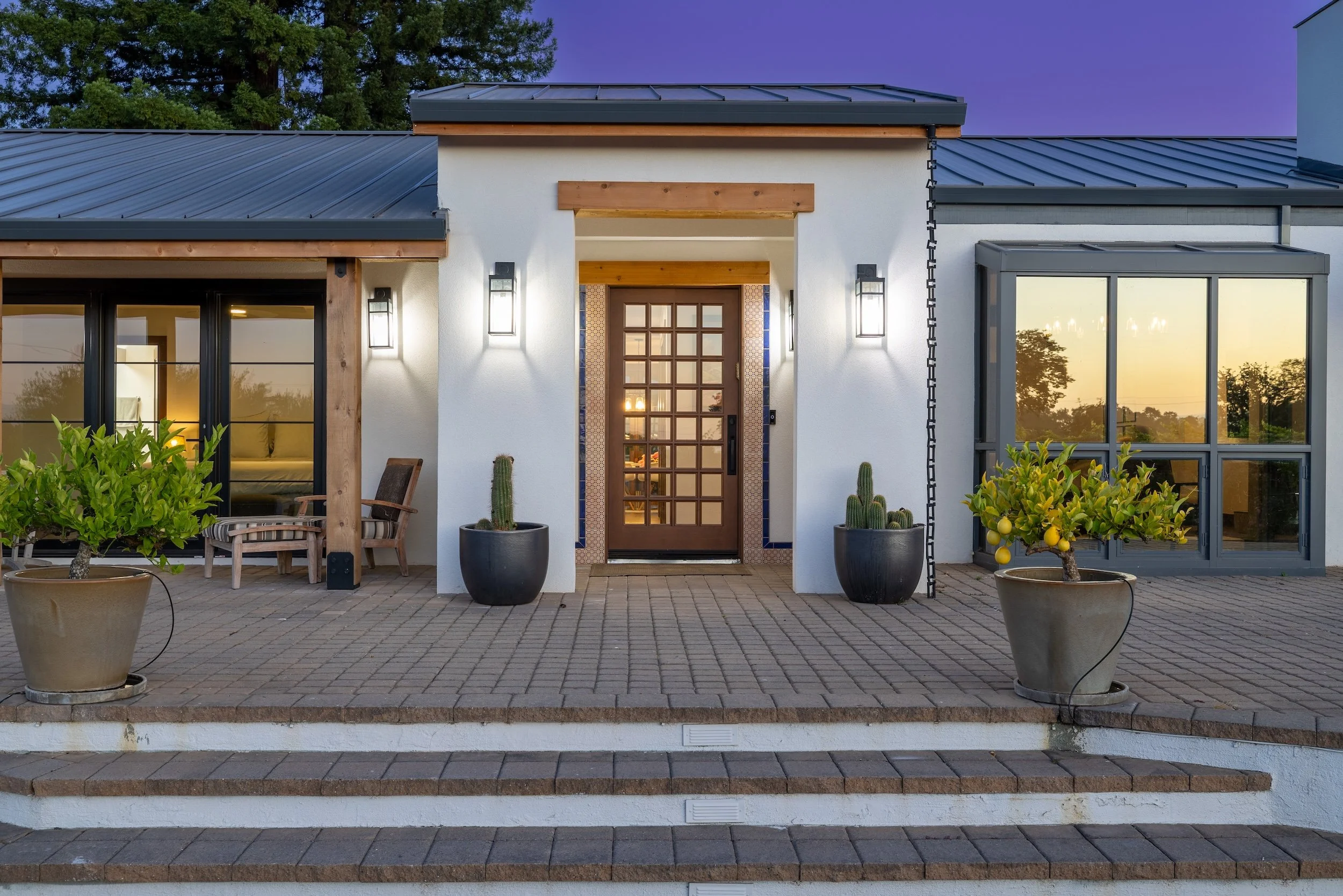 Front view of a modern house with white walls, large windows, and a wooden front door, decorated with potted cacti and plants, during sunset or twilight.