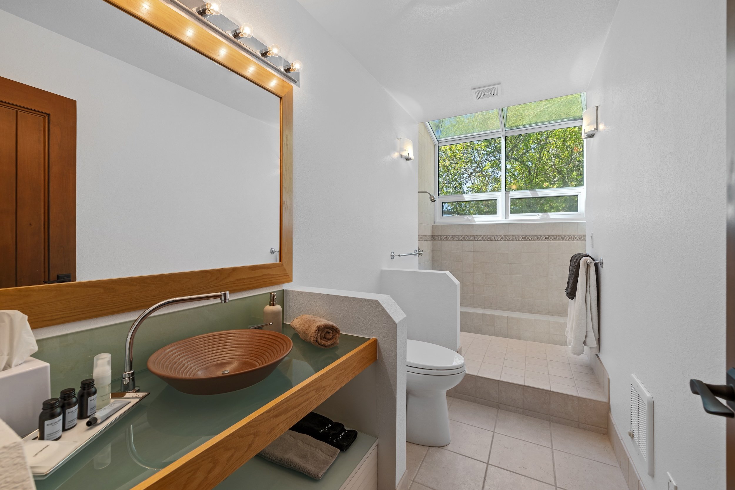 A modern bathroom with white textured walls, a large window with a view of green trees, and a walk-in shower area with beige tiles. There is a wooden vanity with a green countertop, a round copper sink, bottles of toiletries, and a rolled towel. A la