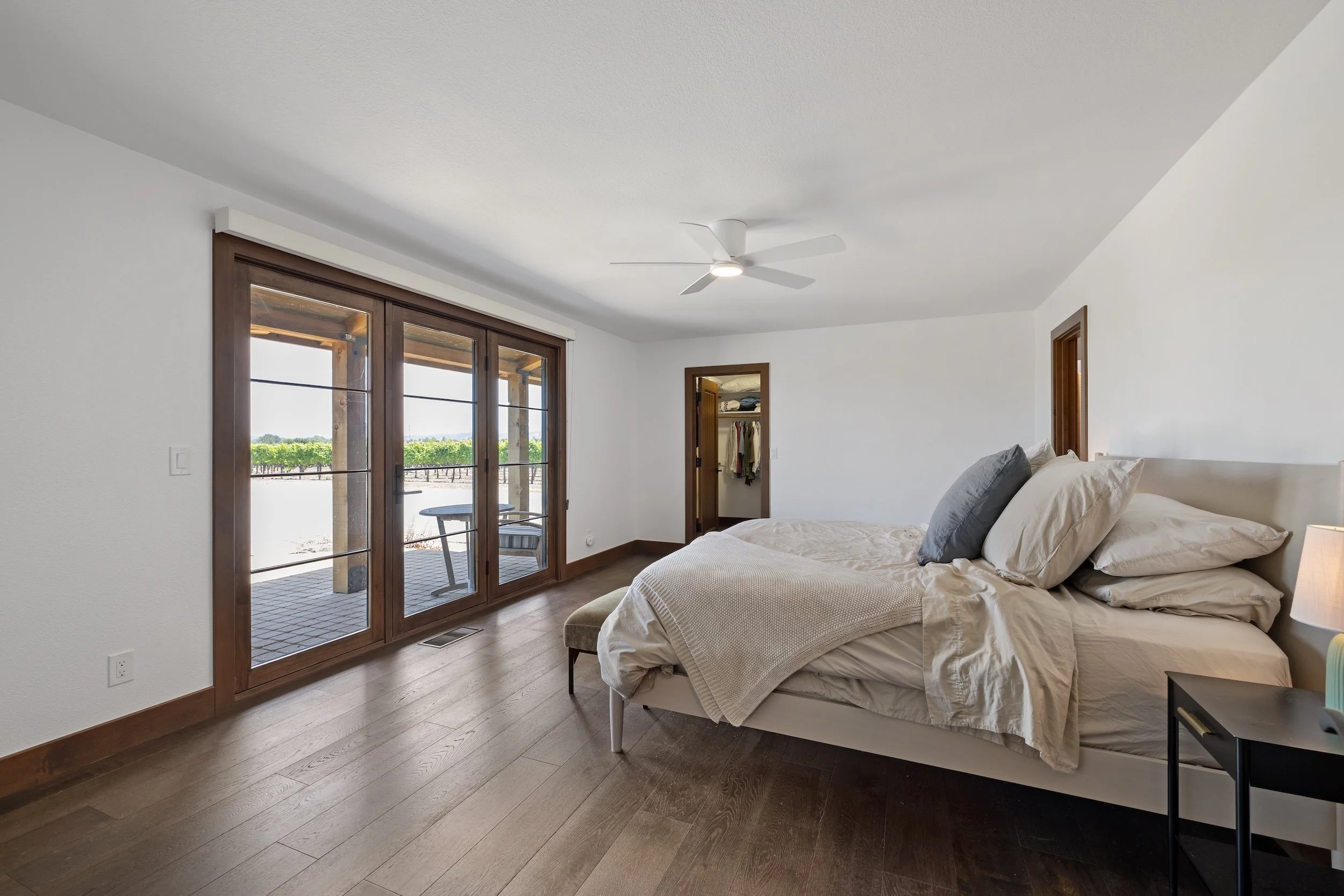 A bedroom with a large bed, pillows, a bedside table, and a ceiling fan. There are glass sliding doors leading to a patio with outdoor furniture, and a view of a vineyard outside.