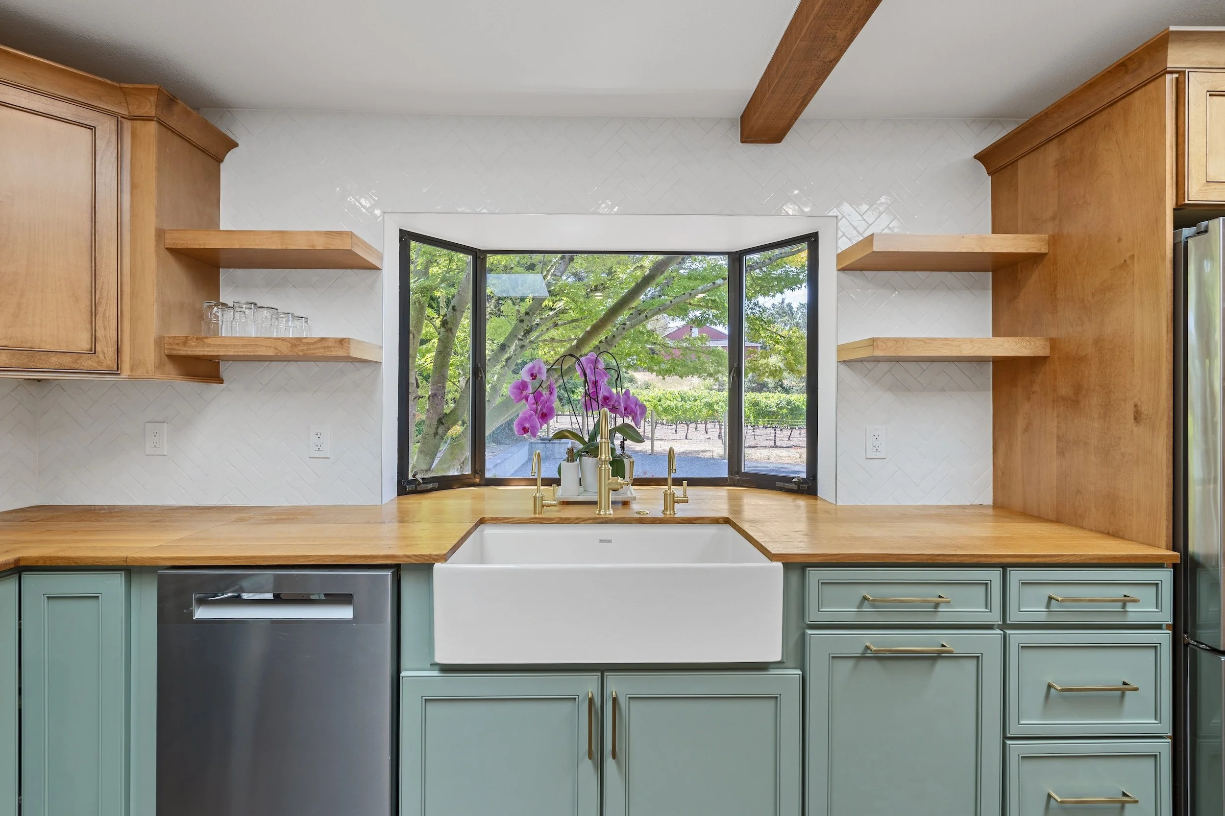 Kitchen with wooden cabinets, large window with a view of trees, and a farmhouse sink with a orchid plant nearby.