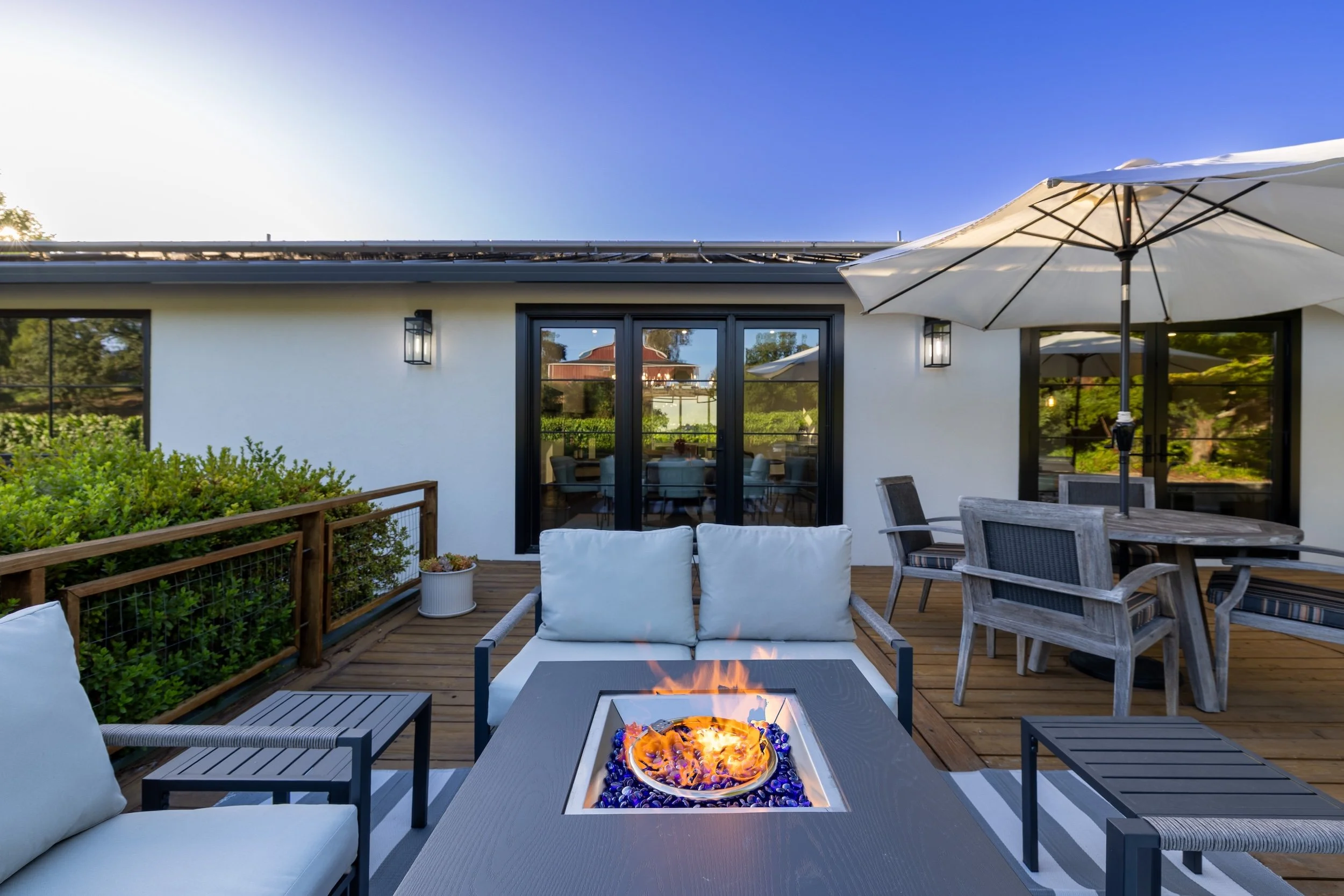 A modern outdoor patio with a fire pit, seating, a table with chairs, an umbrella, and sliding glass doors leading into a house.