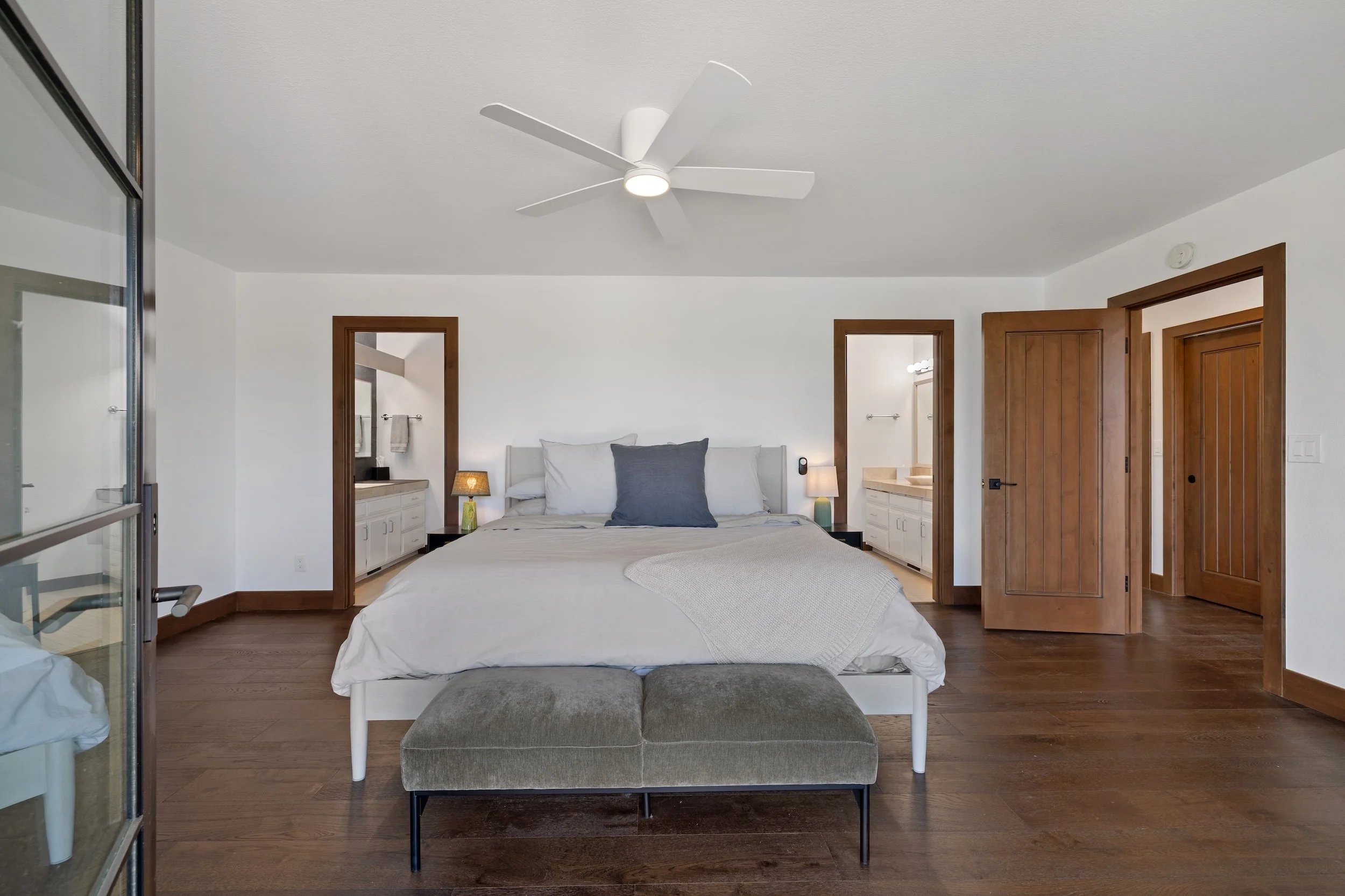 Modern bedroom with a bed, gray blanket, dark blue pillow, two nightstands with lamps, two open doorways, ceiling fan, and wooden flooring.