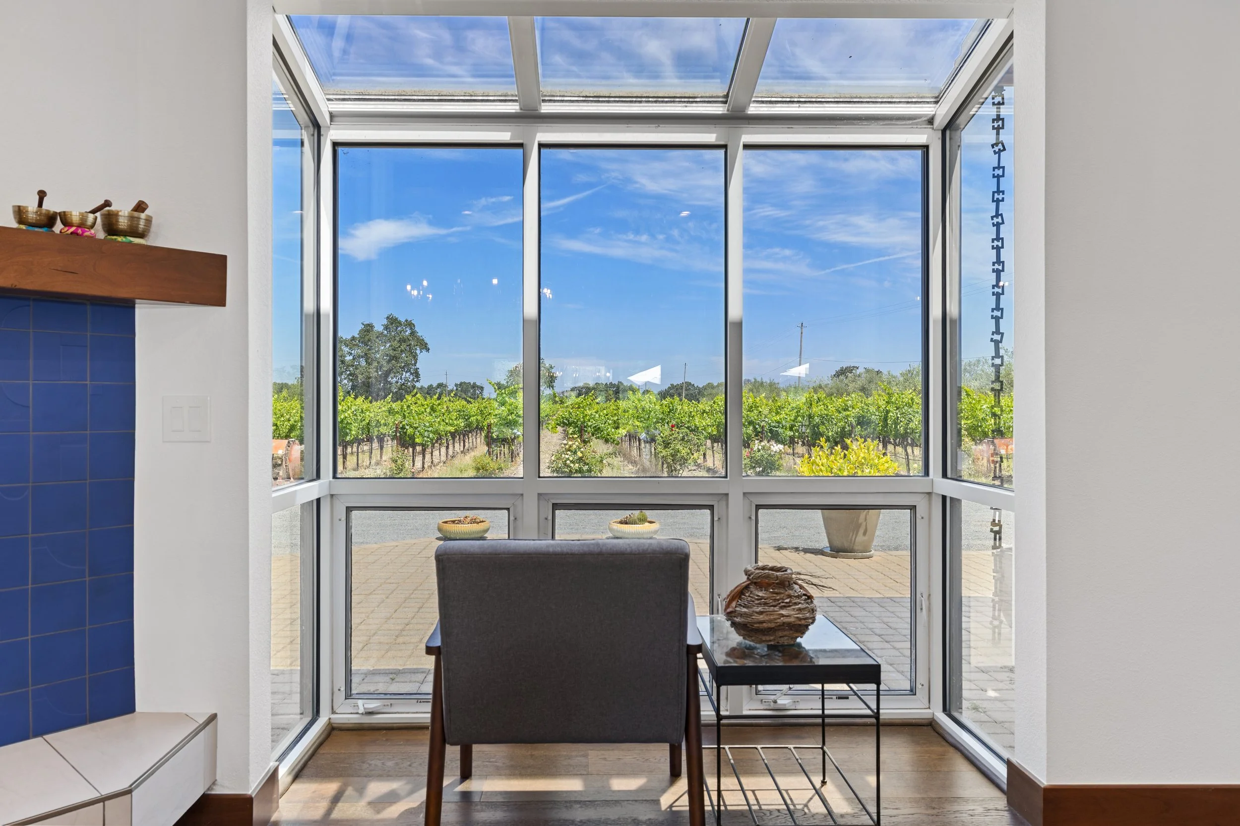 A view through a large glass window with a slanted roof, showing a vineyard and a blue sky with scattered clouds.