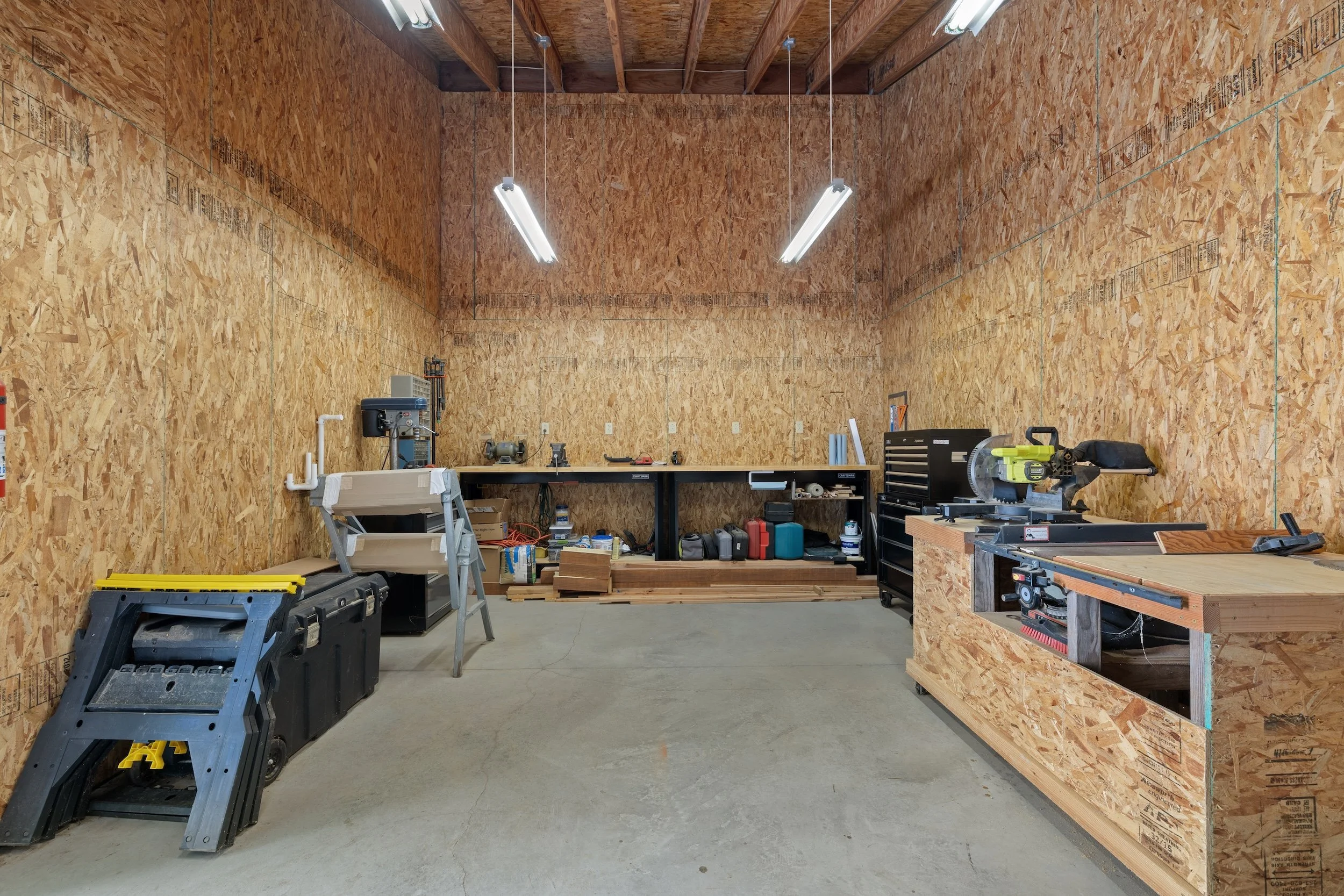 An unfinished workshop with walls and ceiling made of oriented strand board (OSB). There are workbenches, tools, a miter saw, and various supplies organized on shelves and tables. Bright fluorescent lights hang from the ceiling.