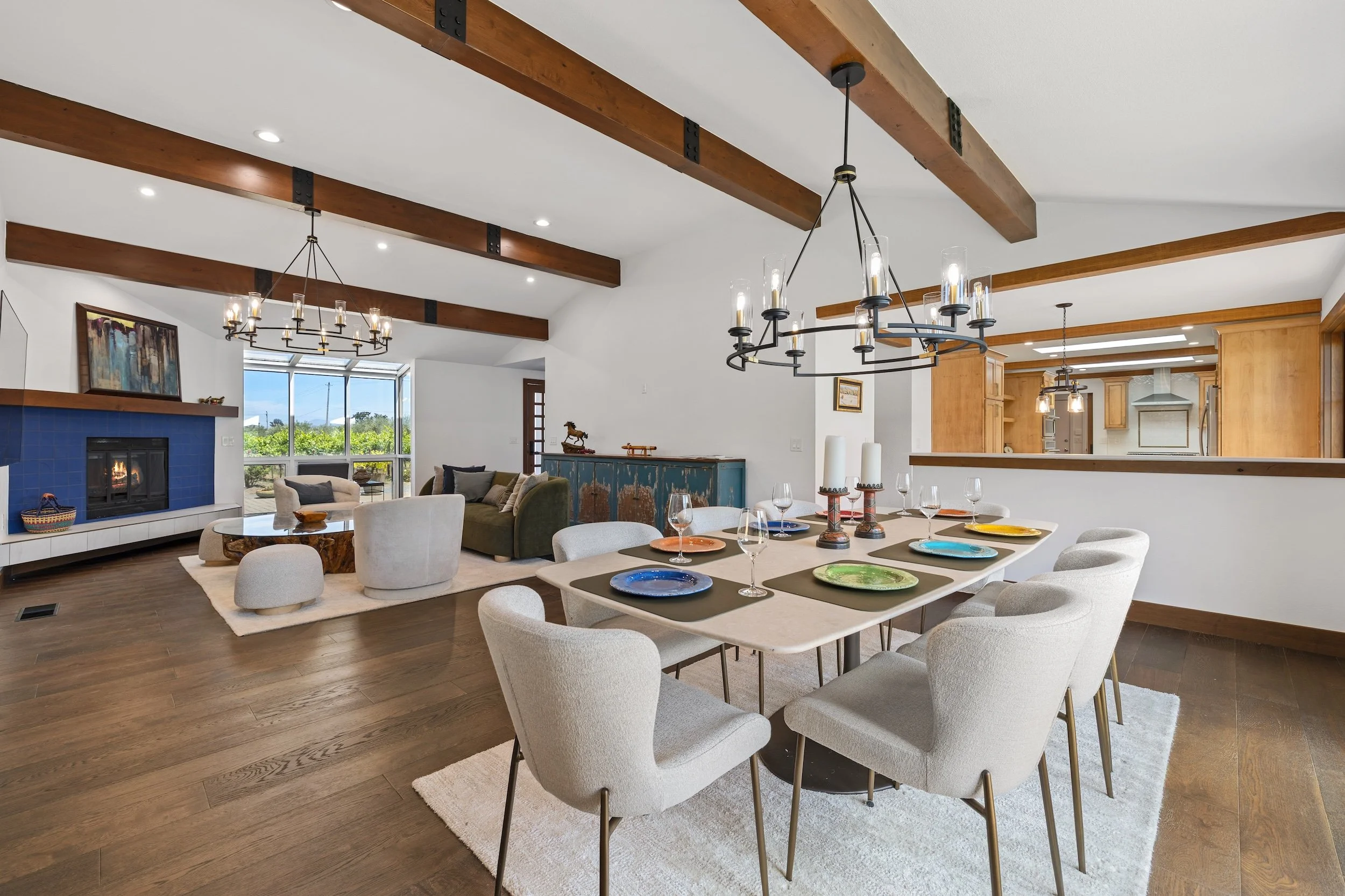 Open-concept living and dining room with wooden beams on ceiling, hardwood floors, a white fireplace with blue tiles, a beige rug, a cozy seating area, a large dining table with colorful plates, and modern chandeliers hanging from the ceiling.