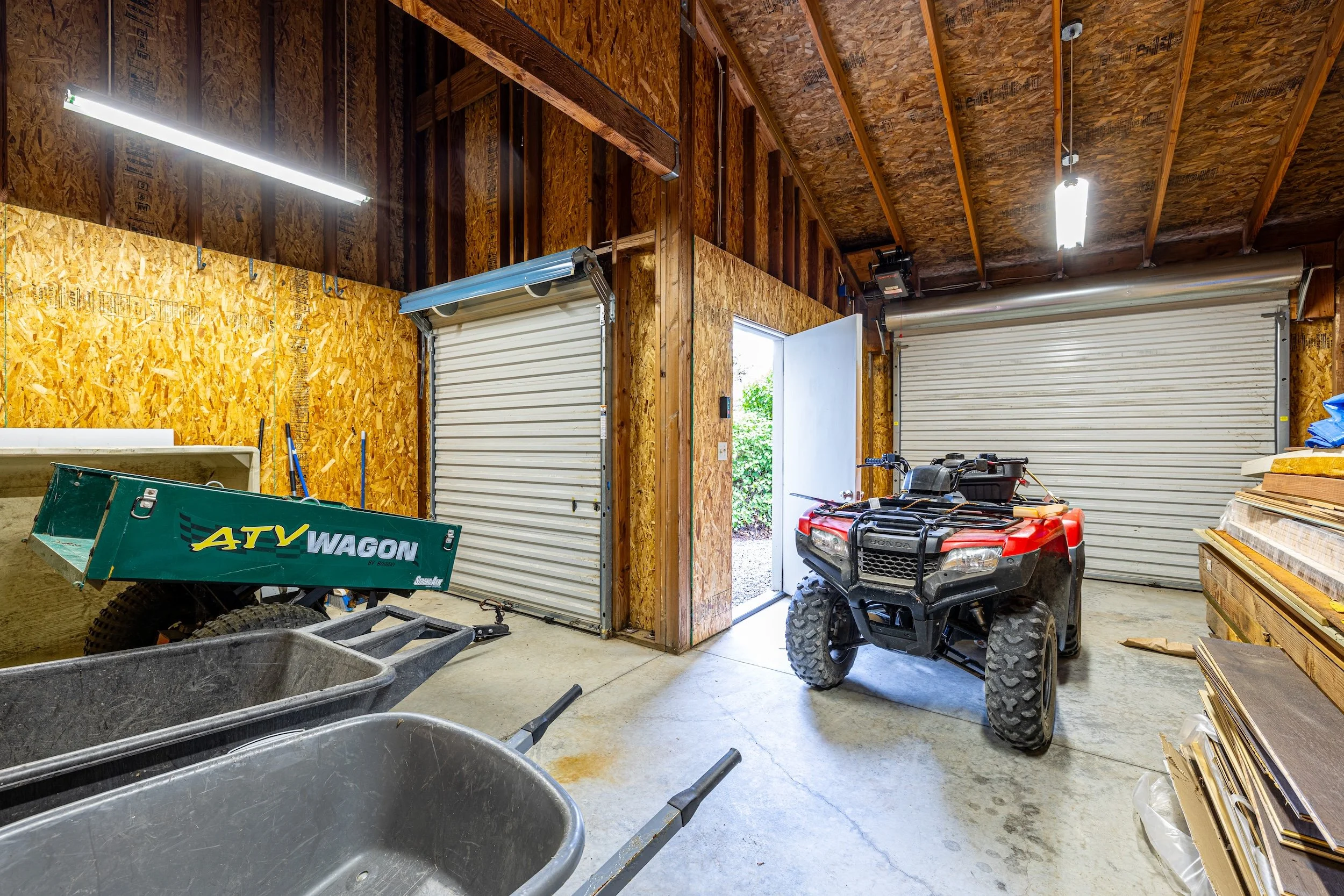 An indoor garage with partially finished walls and ceiling, two open garage doors, a small red ATV, a lawn mower, a green ATV wagon, and various tools and materials scattered around.