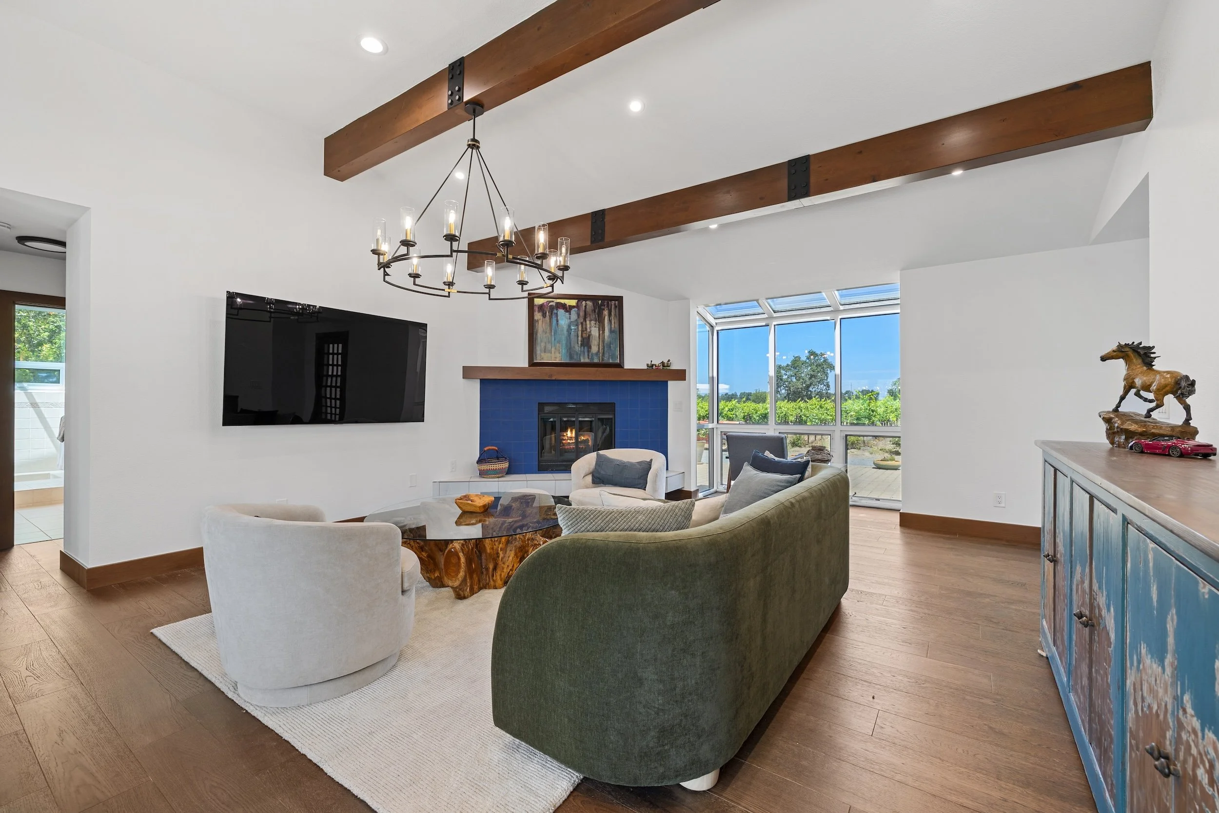 Spacious living room with a white ceiling, wooden beams, a black mounted flat-screen TV, a fireplace, large windows, and various seating including white, green, and beige chairs.