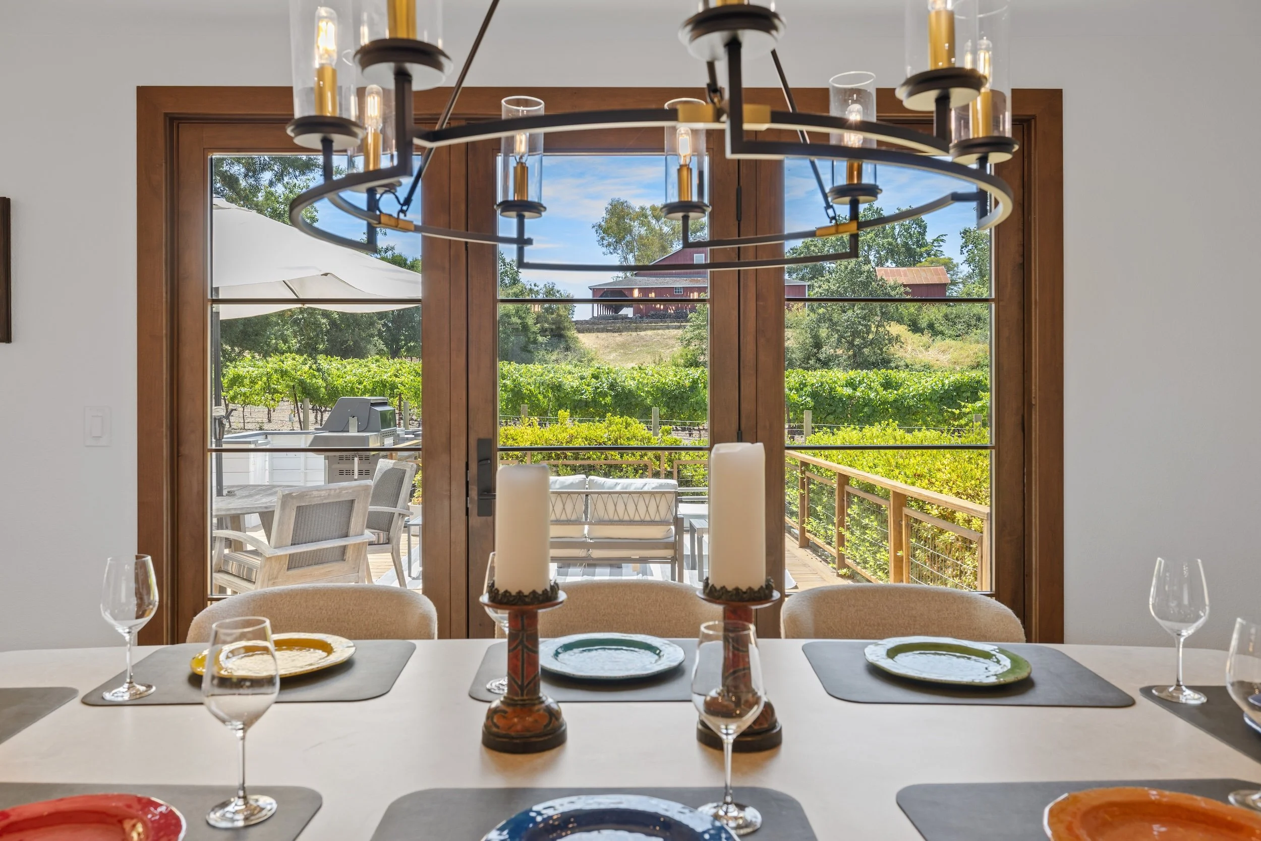 Dining room view through large wooden-framed glass doors shows an outdoor deck with patio furniture, trees, and a red barn in the background, seen during the daytime.