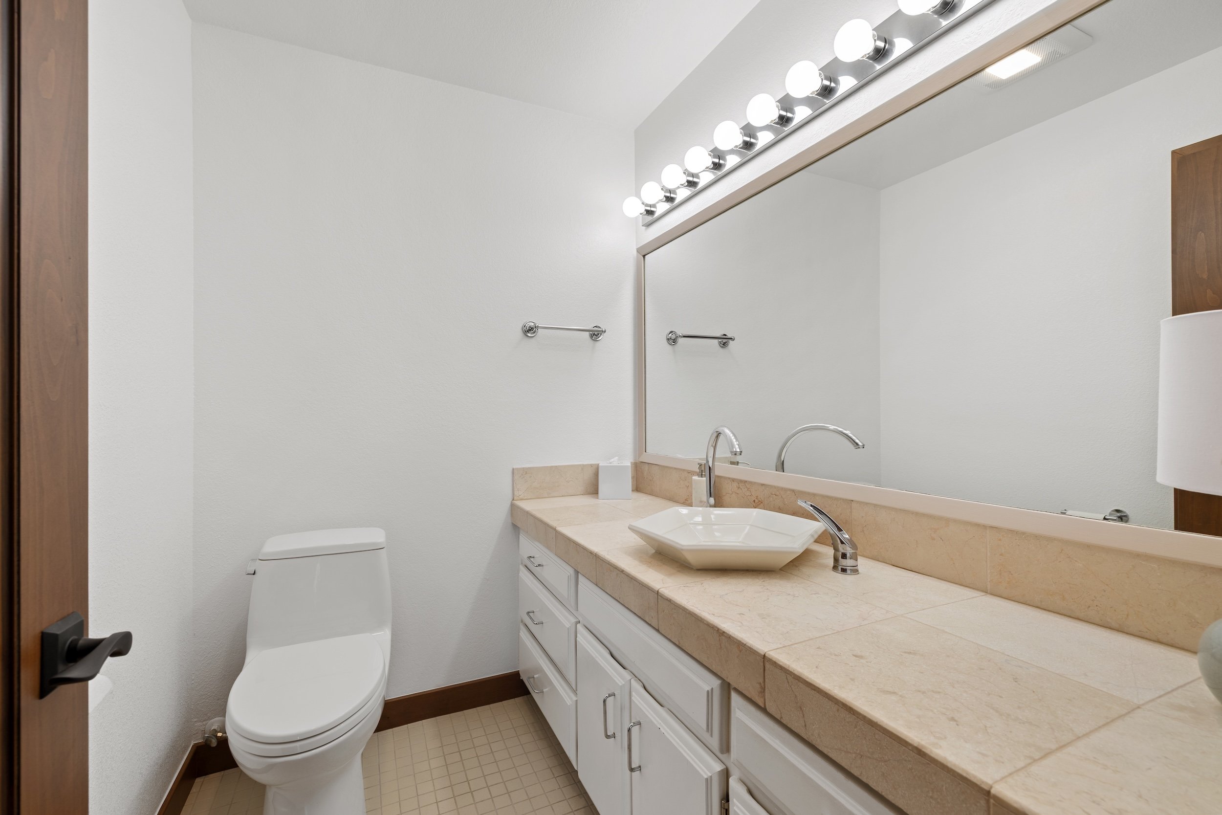 A bathroom with a toilet, a large mirror, a sink with a beige marble countertop, and white cabinetry underneath. The bathroom has white walls, a towel bar, and a row of vanity lights above the mirror.