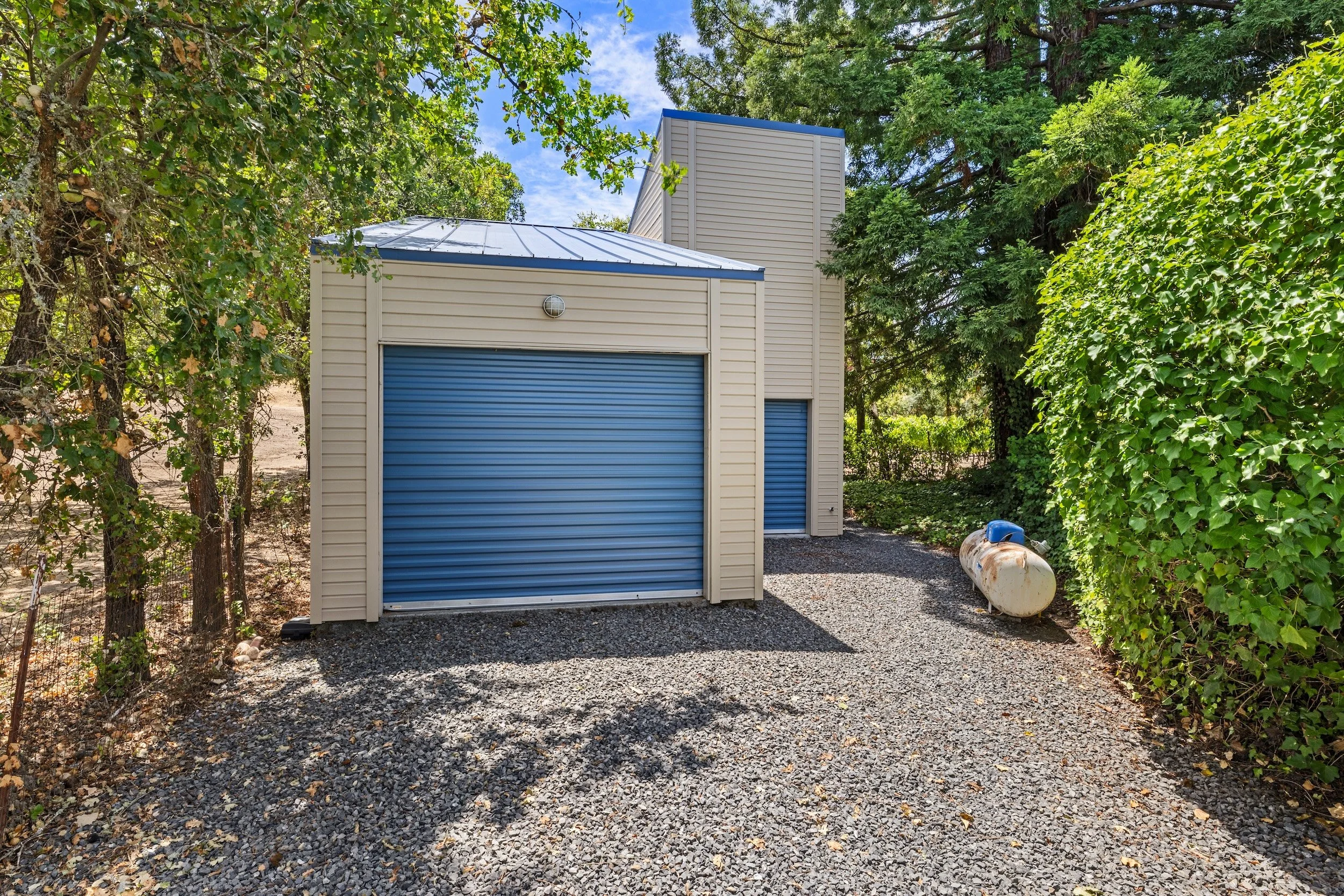 A small beige shed with blue roll-up doors located in a gravel yard, surrounded by tall green trees and bushes, with a propane tank on the right side and a bright blue sky with some clouds overhead.
