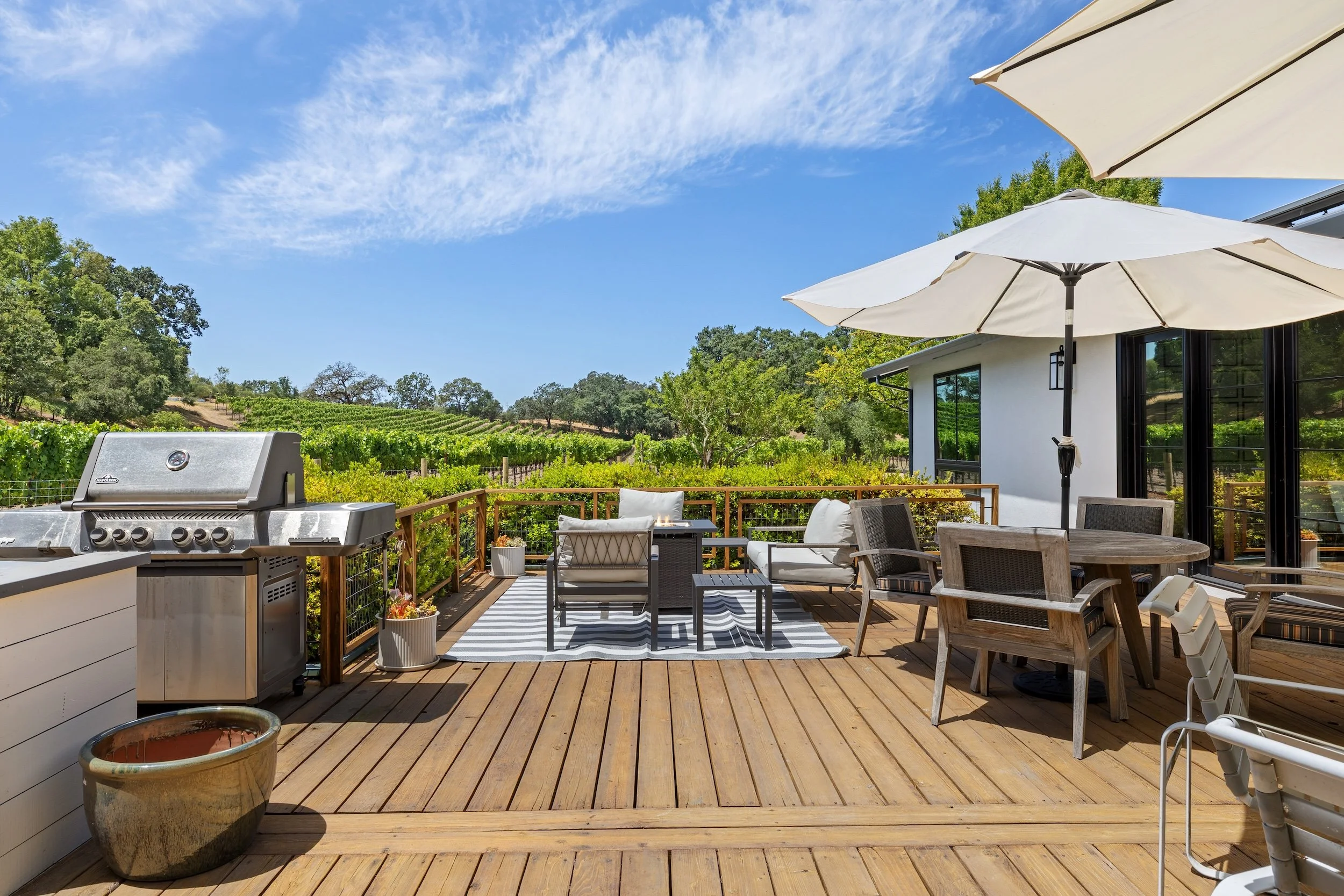 A spacious outdoor patio with wooden flooring, attractive seating, shaded by large white umbrellas, overlooking a scenic vineyard landscape with green trees and rolling hills under a partly cloudy blue sky.