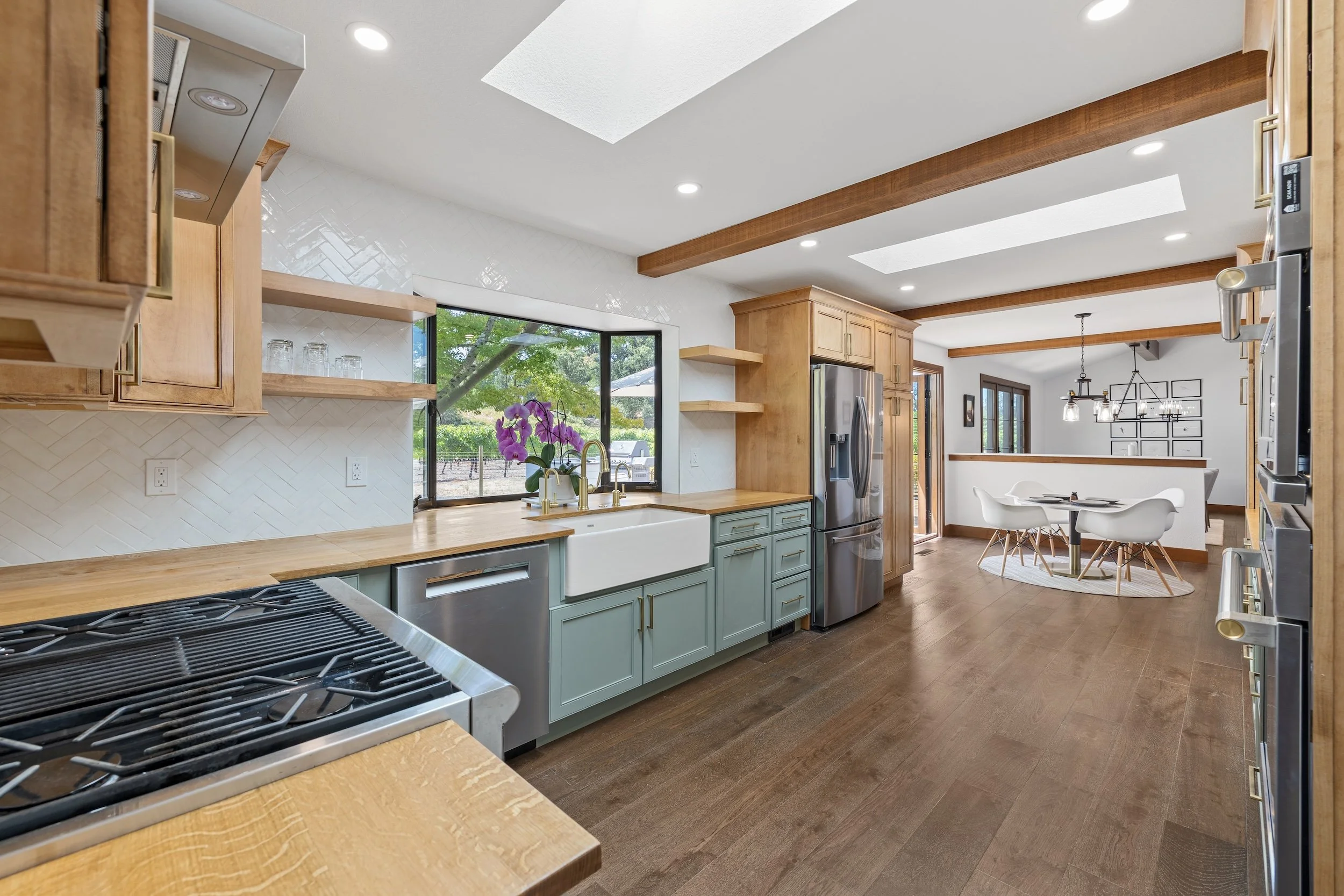 Modern kitchen with wooden cabinets, white walls, green lower cabinets, stainless steel appliances, large window with outdoor view, purple orchids on the counter, and a dining area with white chairs and a round table.