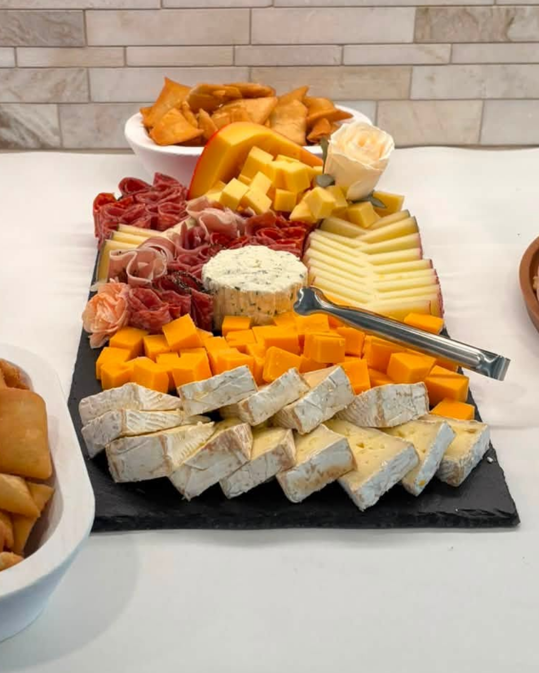 Assorted cheeses and cured meats on a black slate cheese board, with a bowl of chips in the background.