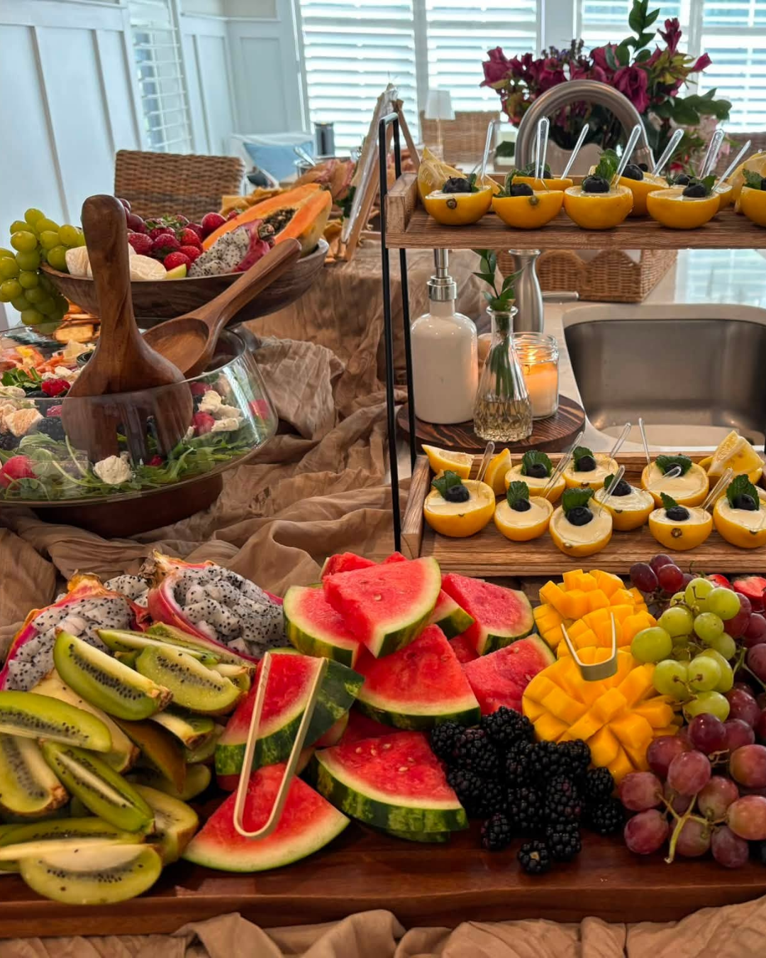 An arranged display of fresh sliced watermelon, mango, grapes, blackberries, kiwi, and dragon fruit on a wooden table, with bowls of berries and lemon slices, set for a fruit buffet.