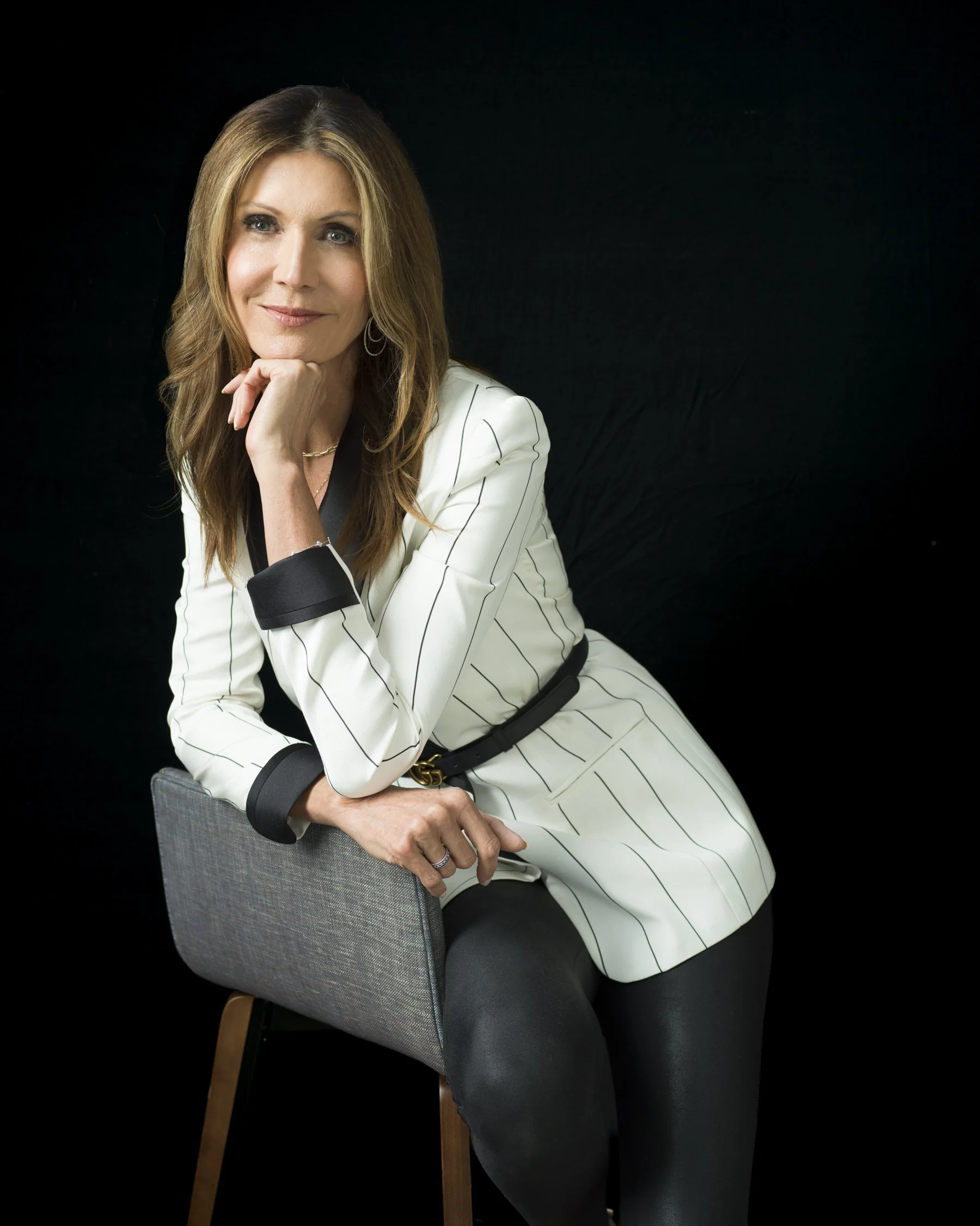A woman with long, light brown hair and blue eyes, wearing a white pinstripe blazer and black pants, is sitting sideways on a gray chair with wooden legs against a black background. She has a gentle smile and rests her chin on her right hand.