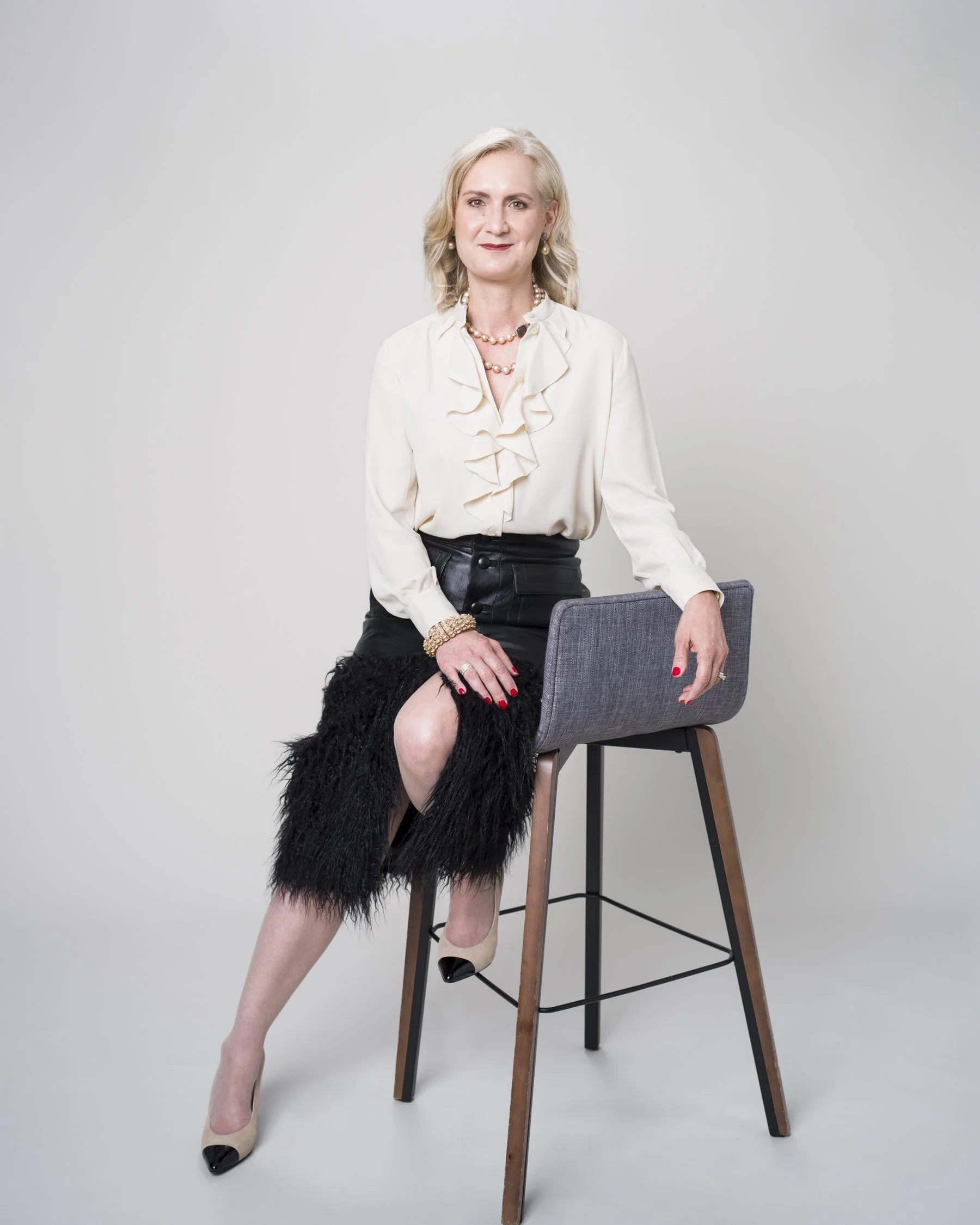 A mature woman with blonde hair dressed in a cream blouse with ruffles, black leather skirt, and beige high heels with black tips, sitting on a tall stool with one leg crossed over the other, against a plain light gray background.