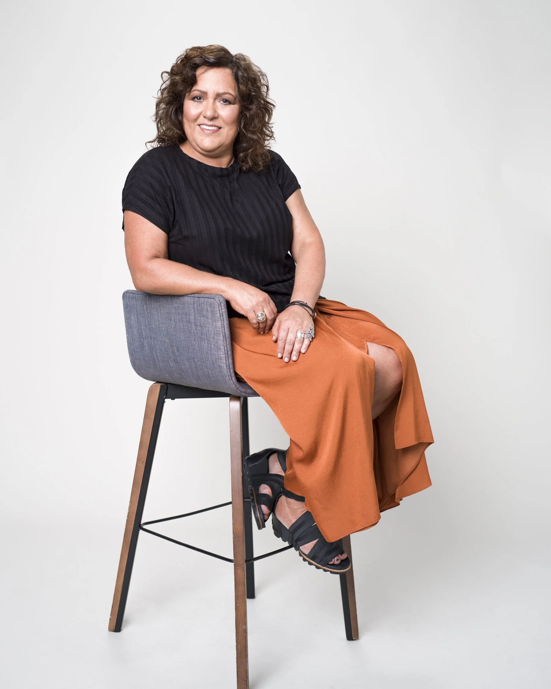 A woman with curly brown hair sitting on a high stool against a plain white background. She is wearing a black top, orange wide-leg pants, and black sandals. She has several rings and bracelets and smiles at the camera.