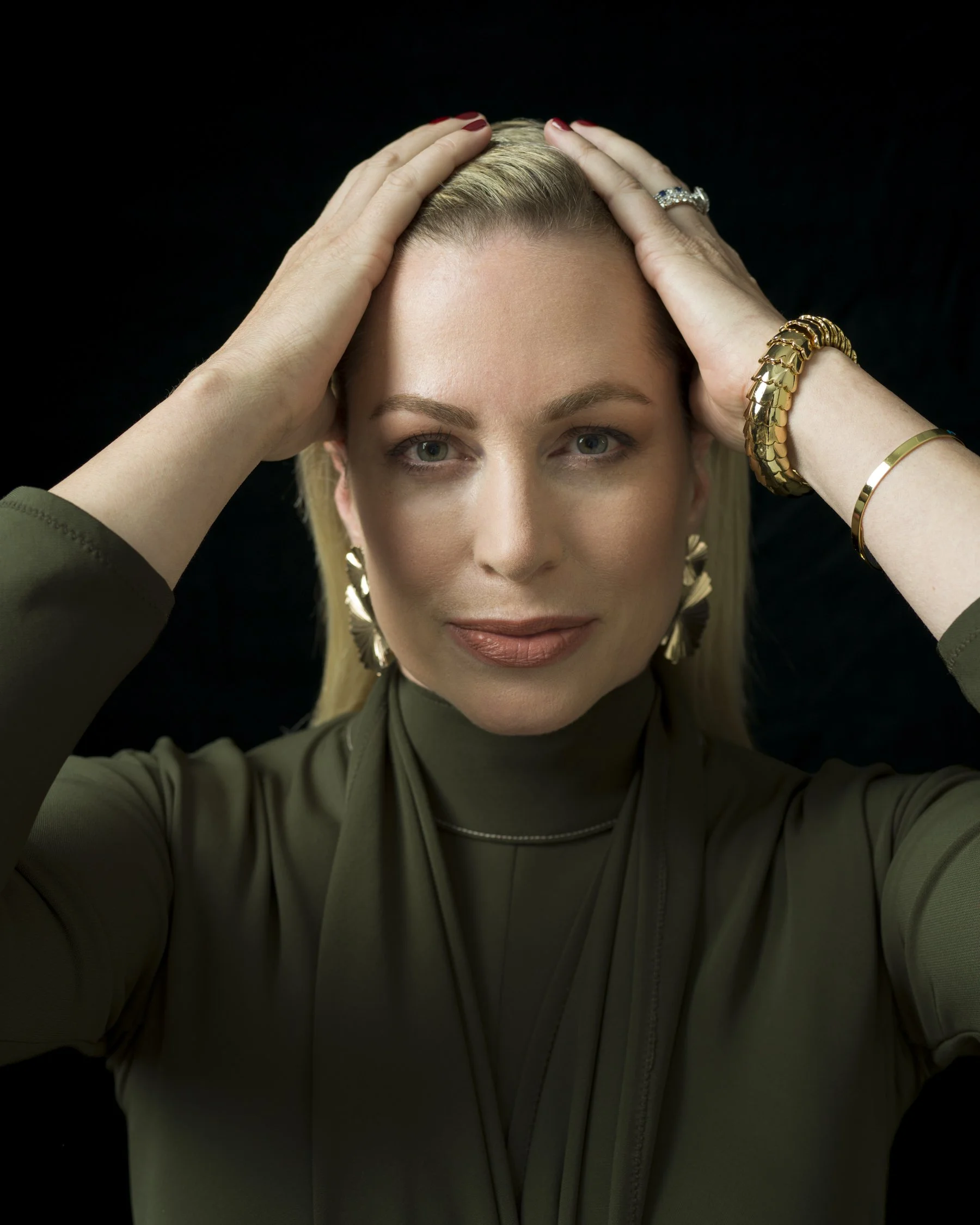 A woman with blonde hair and blue eyes, pressing her hands on her head, wearing rings and bracelets, against a black background.