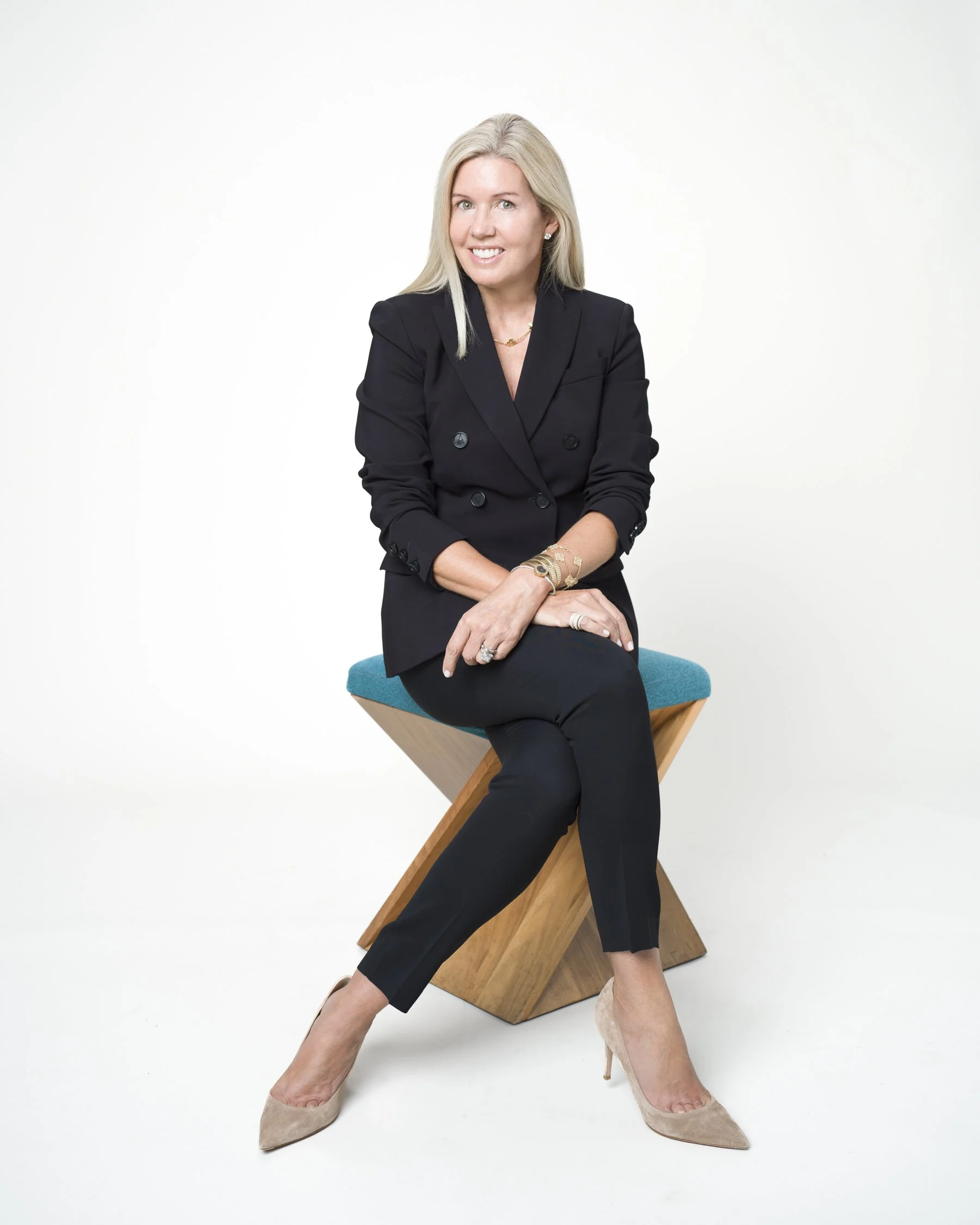 A woman with blonde hair smiling, wearing a black blazer, sitting on a blue and wooden modern chair against a white background, with her legs crossed and wearing beige high heels.
