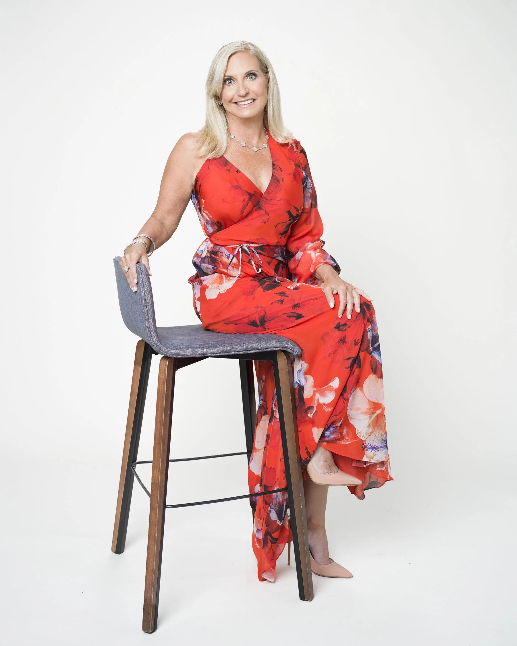 A blonde woman in a red floral dress sitting on a stool, smiling, with an empty white background.