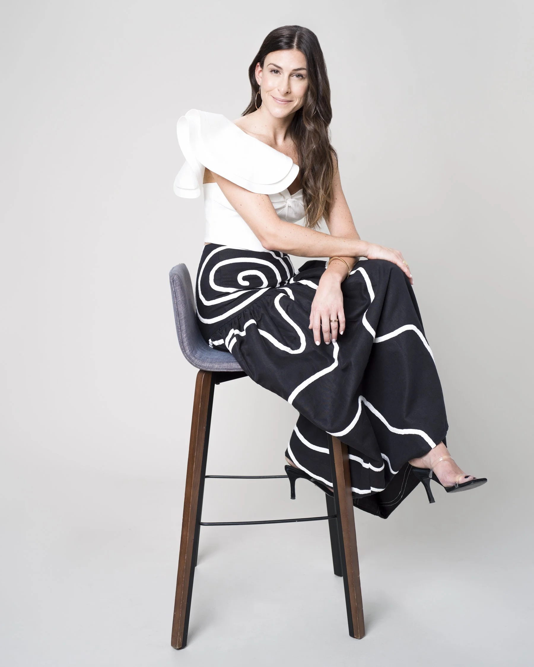 A woman with long brown hair sitting on a wooden stool with a gray seat against a light gray background. She is wearing a white top with a large shoulder ruffle, black pants with white swirl patterns, open-toed heels, and gold hoop earrings. Her righ