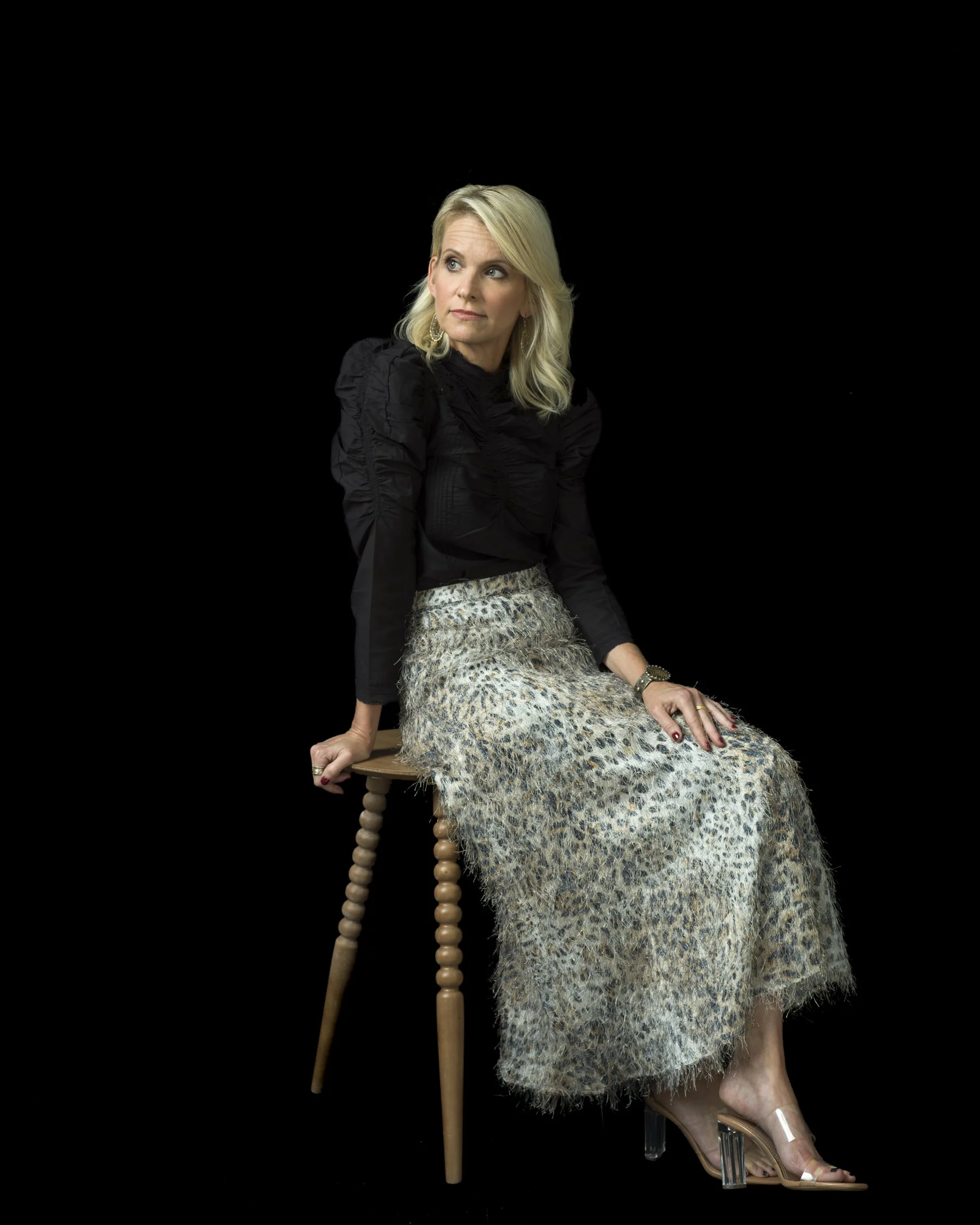 A woman with blonde hair wearing a black top, leopard print skirt, and open-toed heels sitting on a wooden stool against a black background.