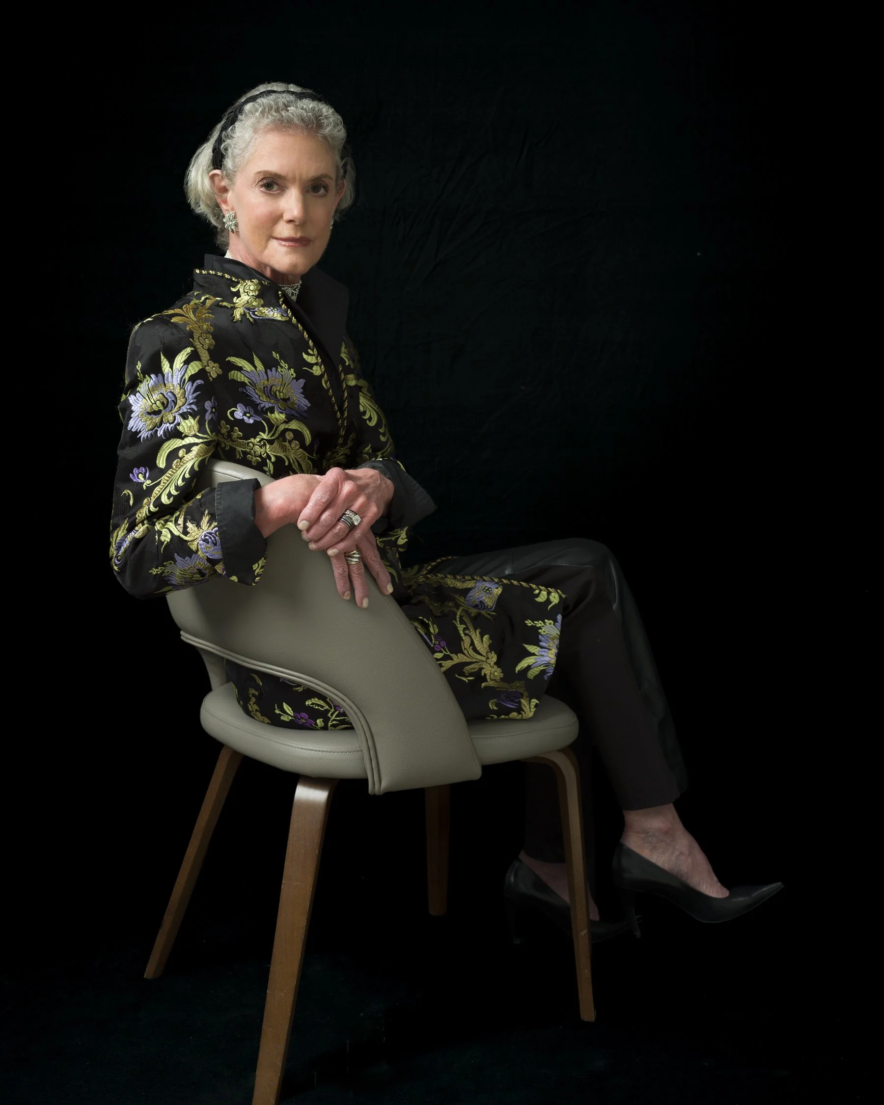 An elderly woman with short gray hair seated on a light-colored chair with wooden legs, wearing black shoes with pink accents, a dark floral jacket, a necklace, earrings, and rings, holding a small object in her right hand against a black background.