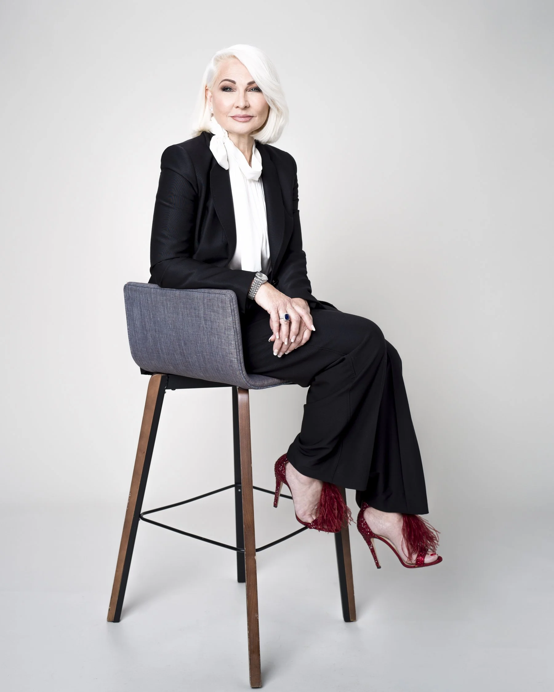 A woman with white hair sits on a high wooden stool in front of a plain white background. She wears a black blazer, white blouse, black pants, and red high-heeled shoes with feathers. She looks at the camera with a slight smile, with her right hand r